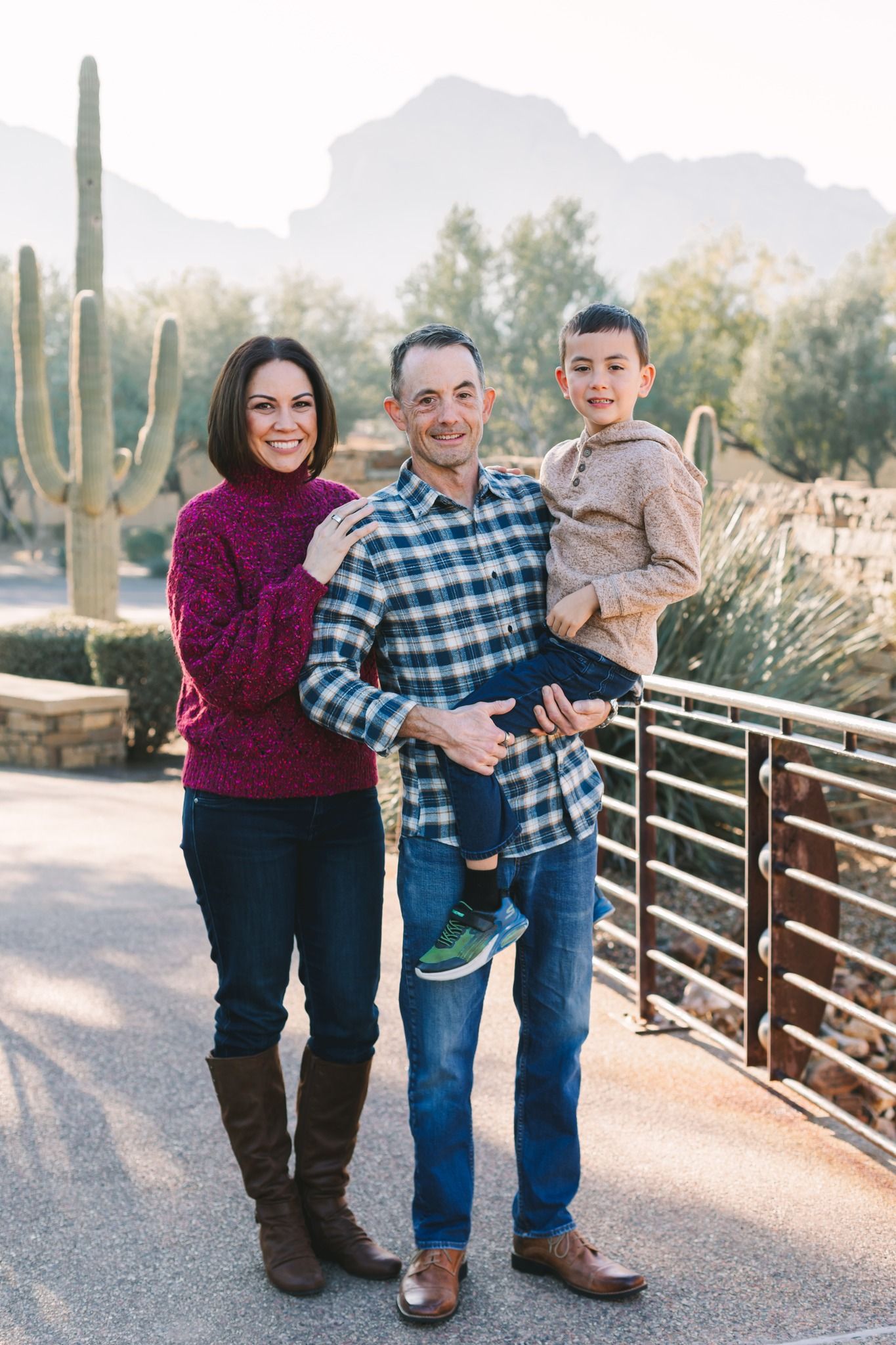Smiling family of three posing on bridge with saguaro cacti and mountains behind them.
