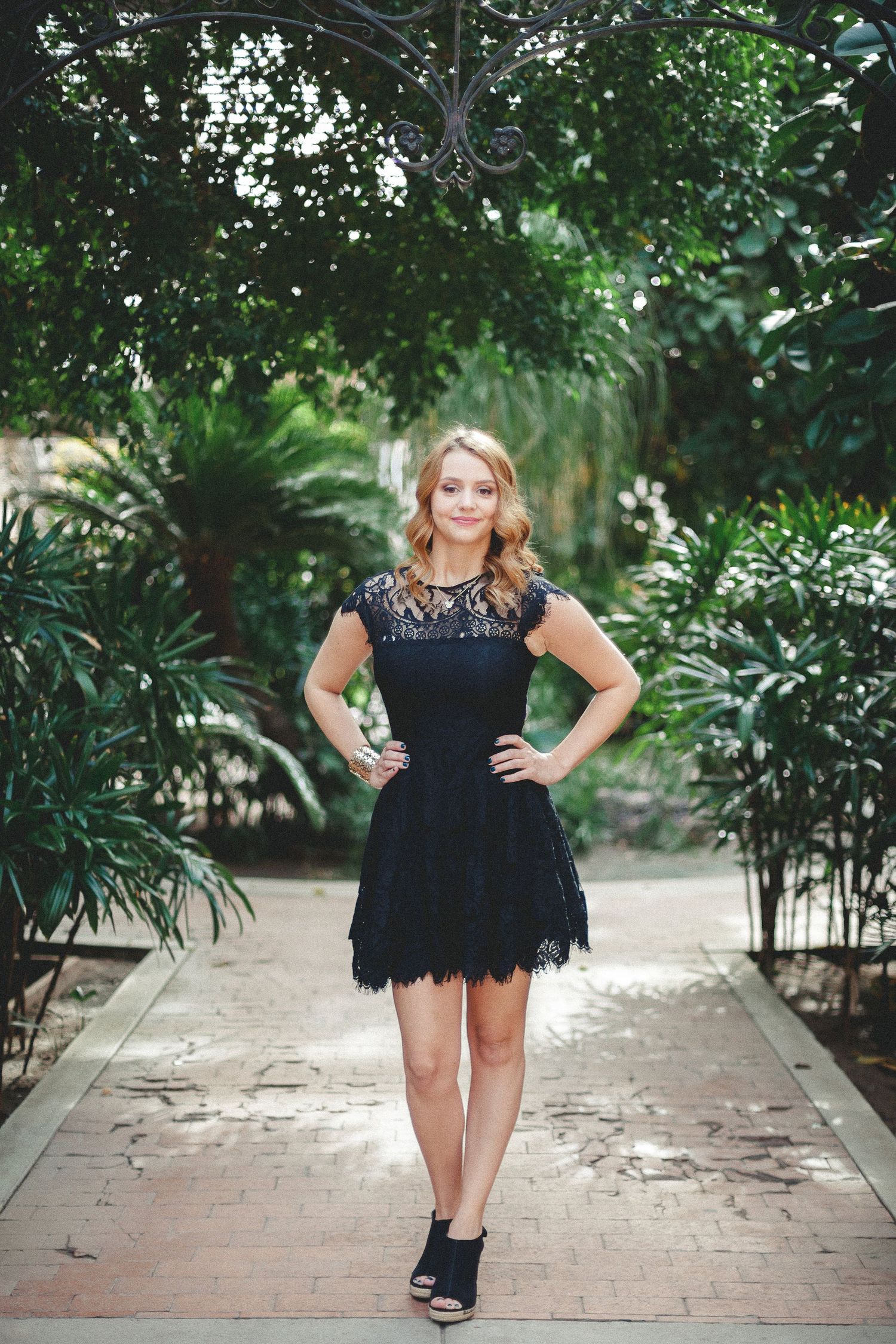 Smiling blonde woman in black lace mini dress standing on garden pathway surrounded by greenery.