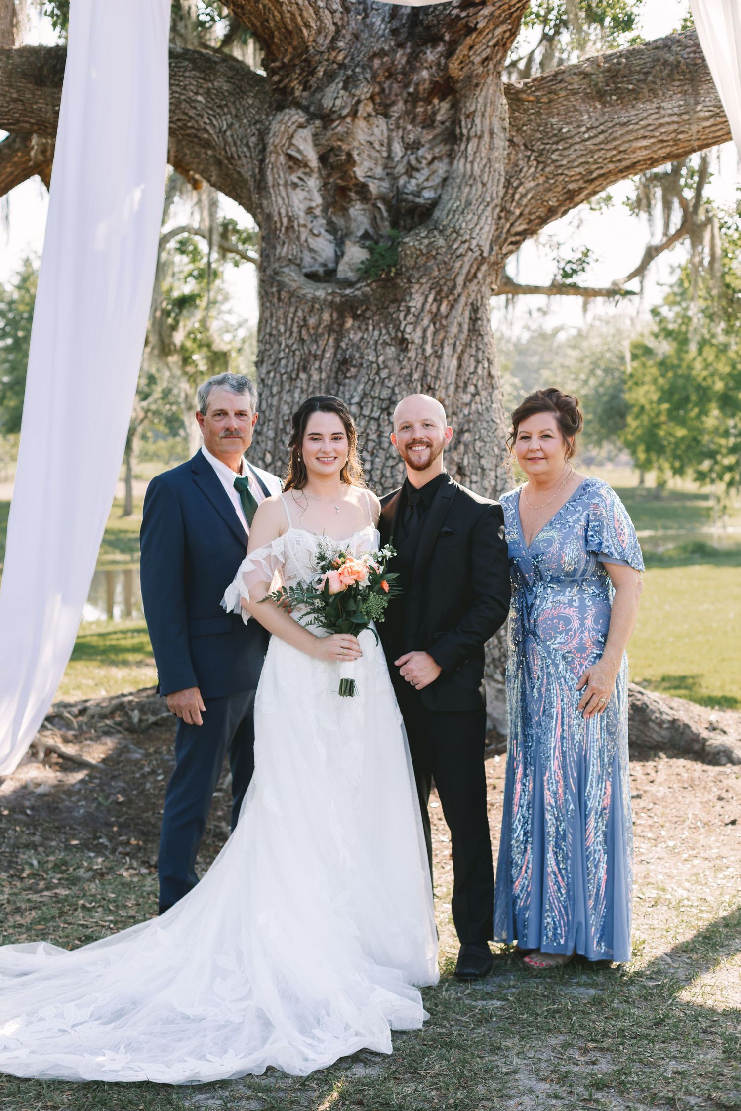 Bride and groom pose with family under a large oak tree at an outdoor wedding ceremony.