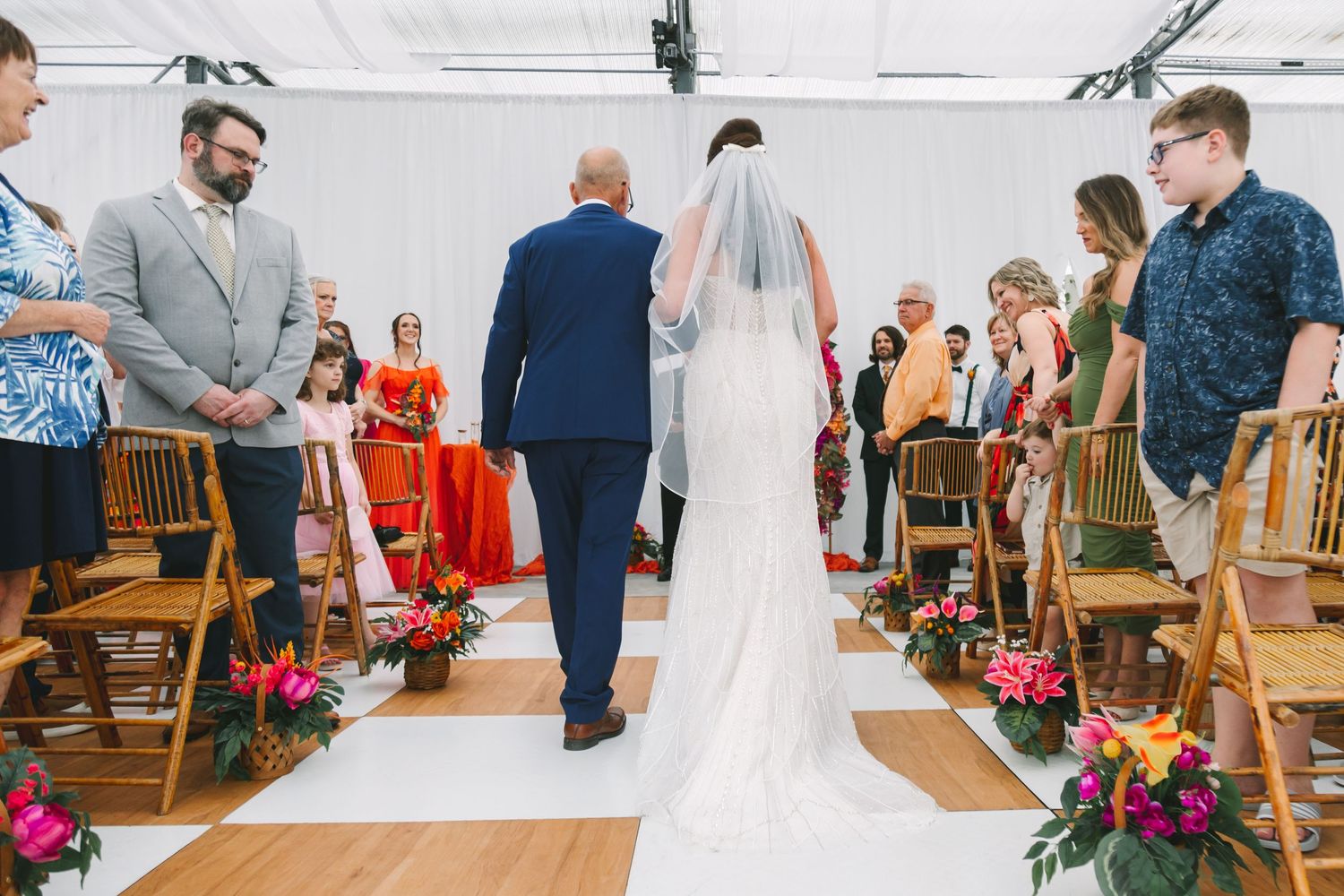 Bride walks down the aisle with escort at outdoor wedding ceremony with colorful floral arrangements.
