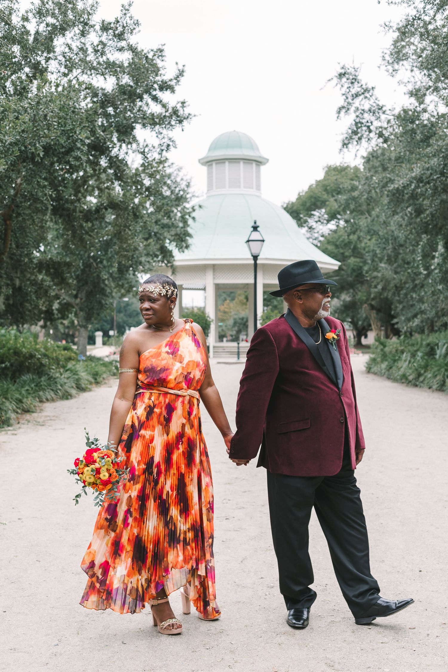 Couple walking hand in hand in colorful attire past a gazebo in a scenic tree-lined park setting.
