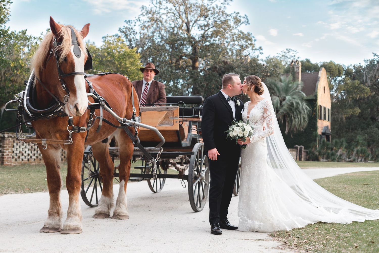 Bride and groom kissing in front of a horse-drawn carriage at an outdoor wedding venue.