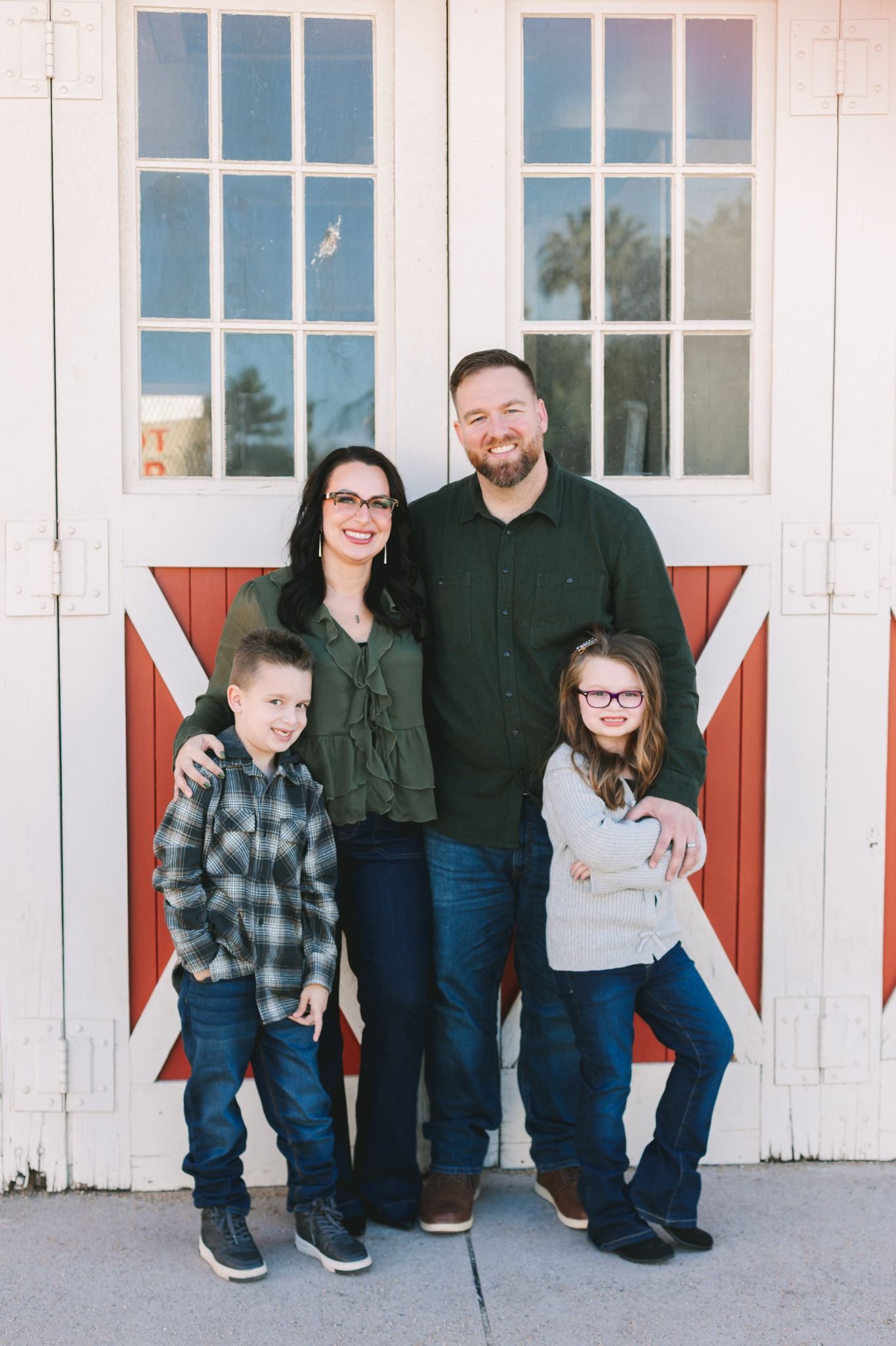 Family of four smiling in front of a red barn door, wearing casual fall outfits.