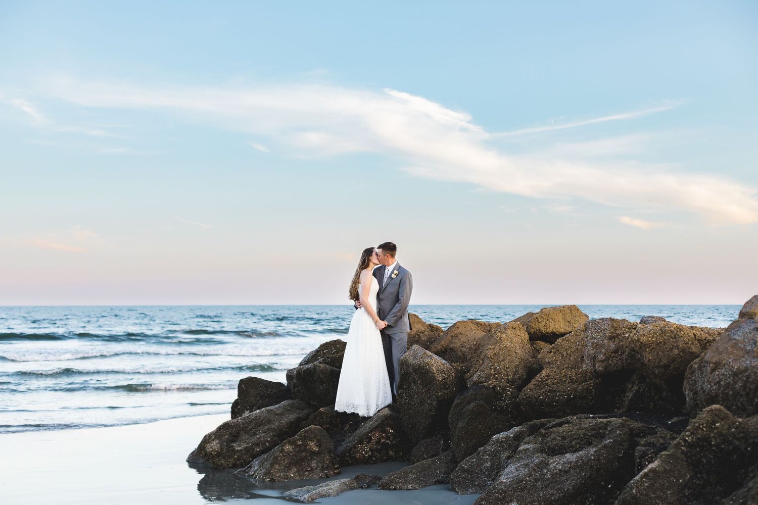 Romantic bride and groom kissing on rocky shoreline with ocean waves and pastel sunset sky behind them.