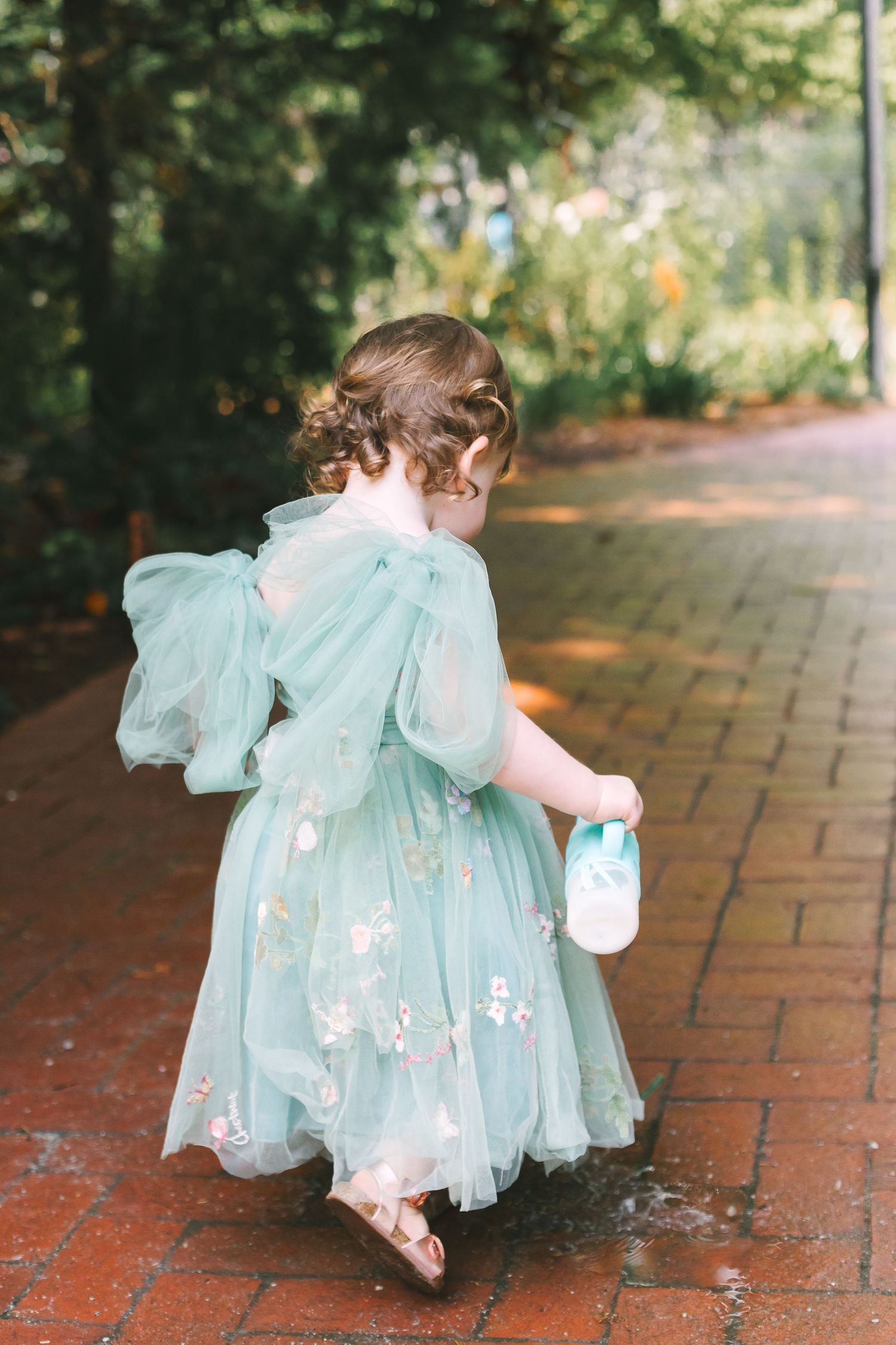 Young flower girl in teal tulle dress walking down brick garden path holding jar.