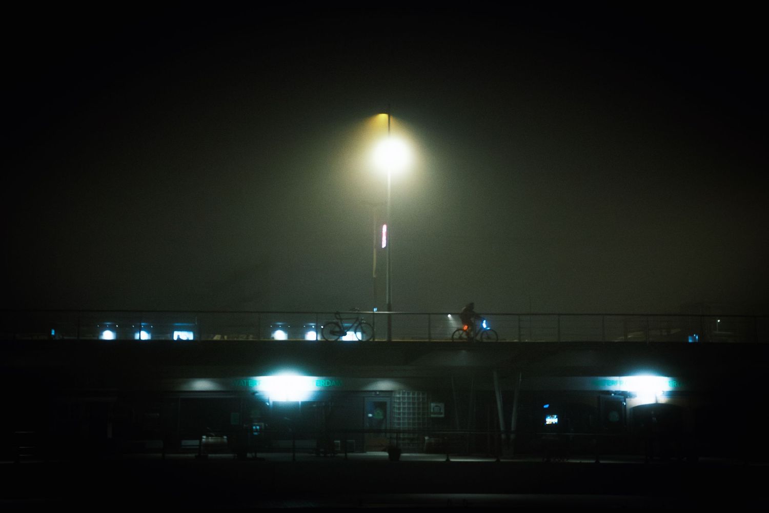 Night street photography in Amsterdam showing the city in heavy mist with soft lights, reflections and a cinematic urban atmosphere.