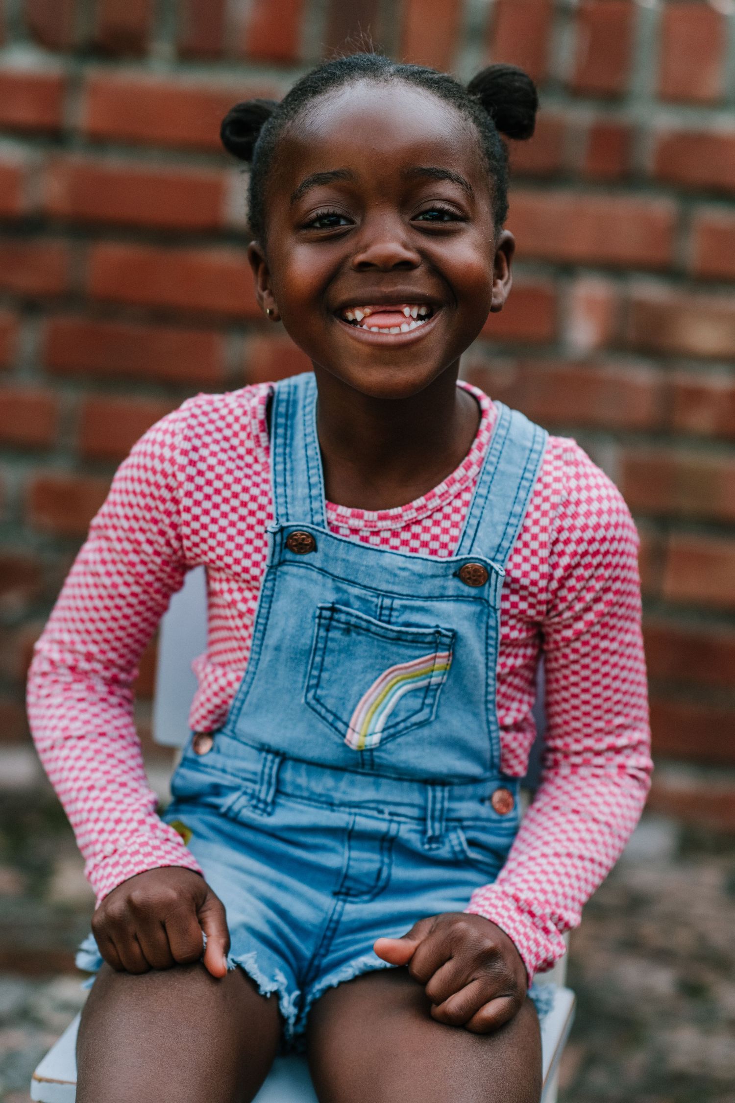 Playful poses in pink gingham top and denim overalls against brick wall.