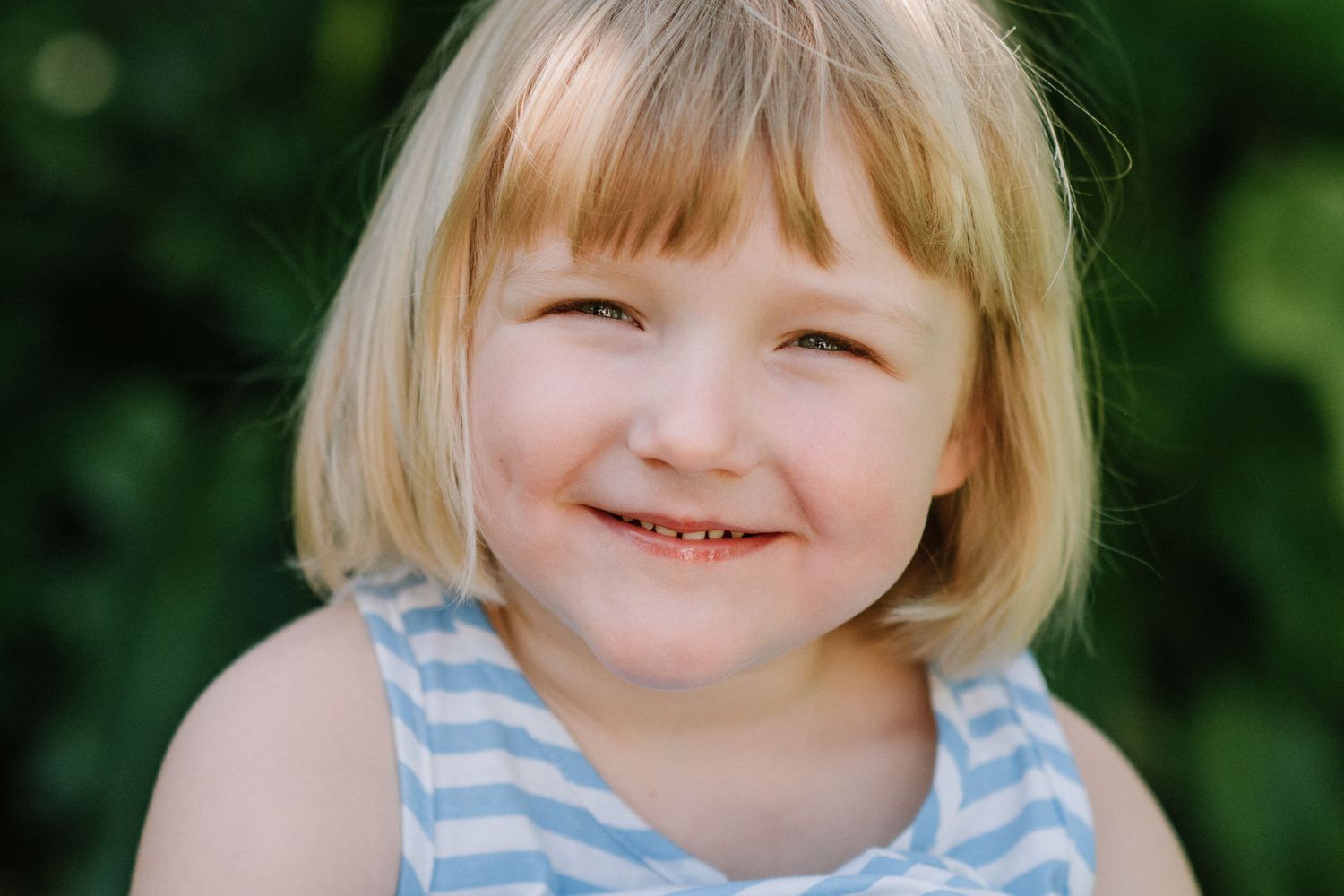 Child in blue striped outfit with short blonde hair smiling at camera outdoors.