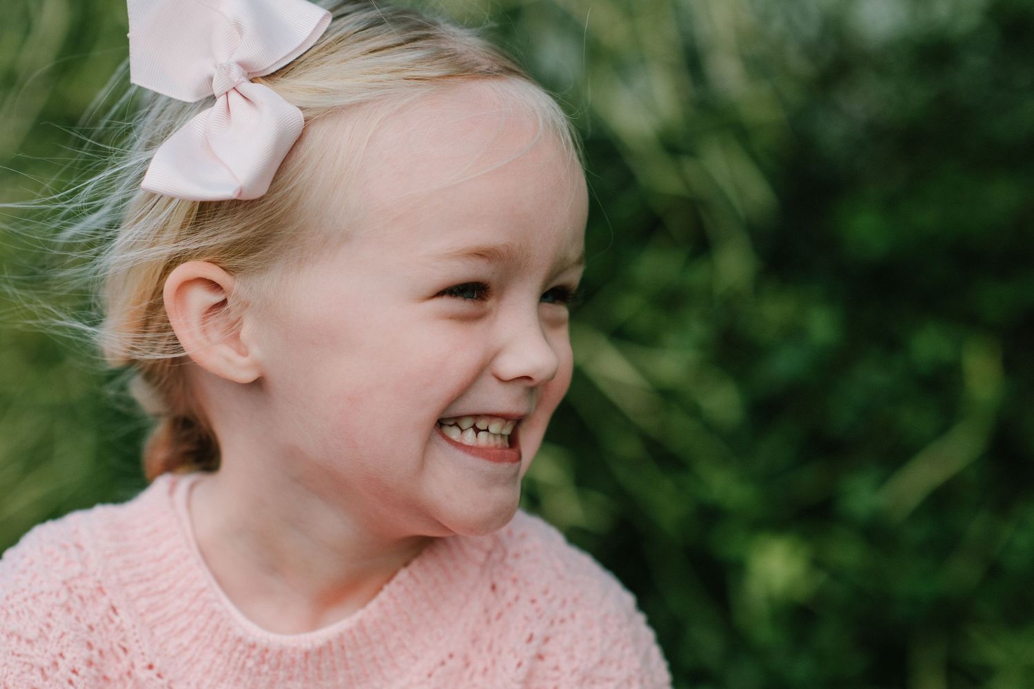 Child in pink sweater with hair bow smiles cheerfully against green outdoor background.