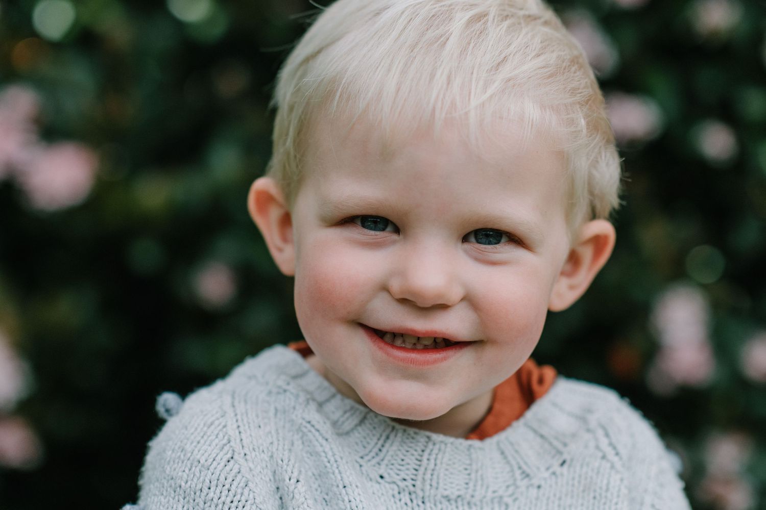 Close-up portrait of a child wearing a grey sweater against a garden background.