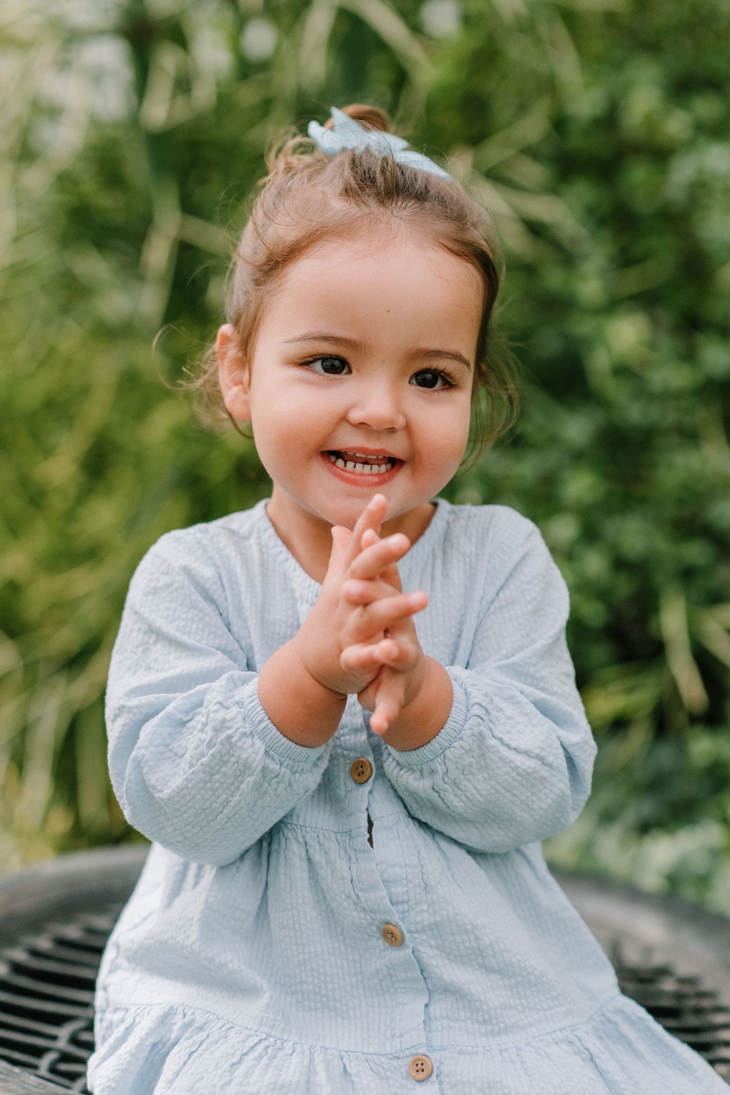 Child in blue dress claps hands while smiling outdoors against greenery.