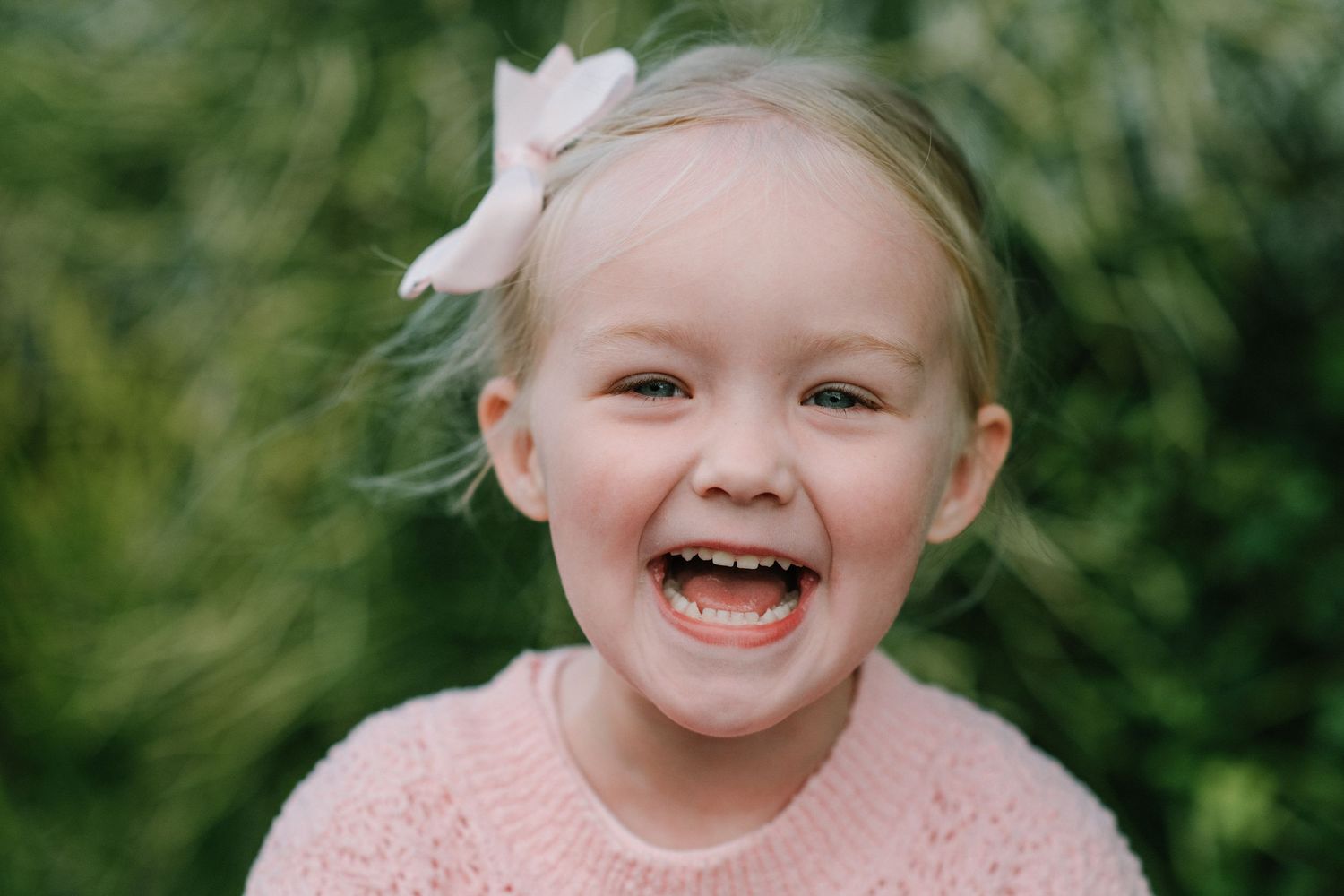 Young child in pink sweater with bow accessory laughs joyfully against green foliage.