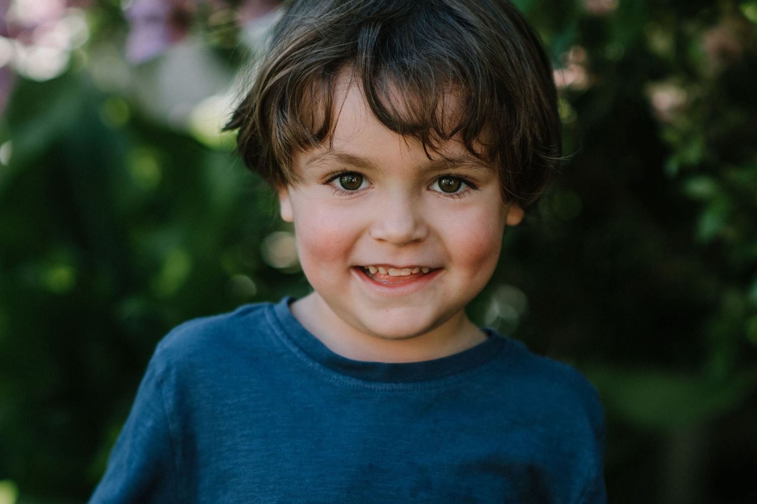 Portrait of a child wearing a blue shirt smiling warmly against a garden background.