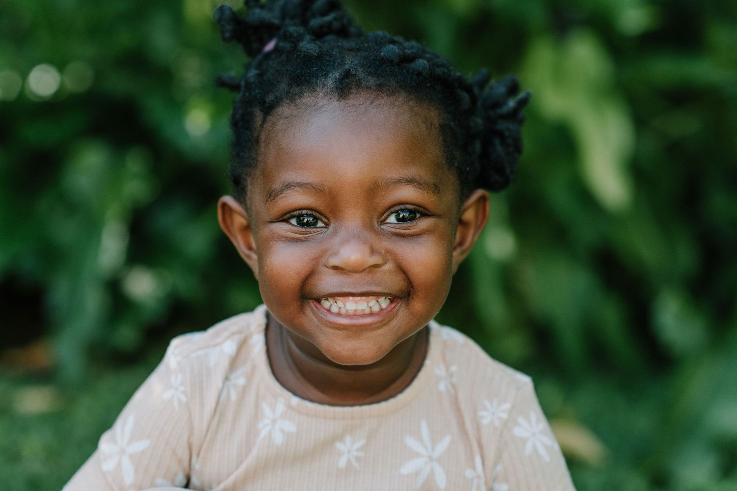 Child in white dress smiling brightly with natural greenery backdrop.