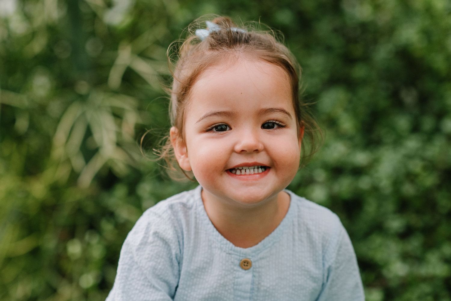 Child in light blue cardigan shows bright smile while sitting outdoors.