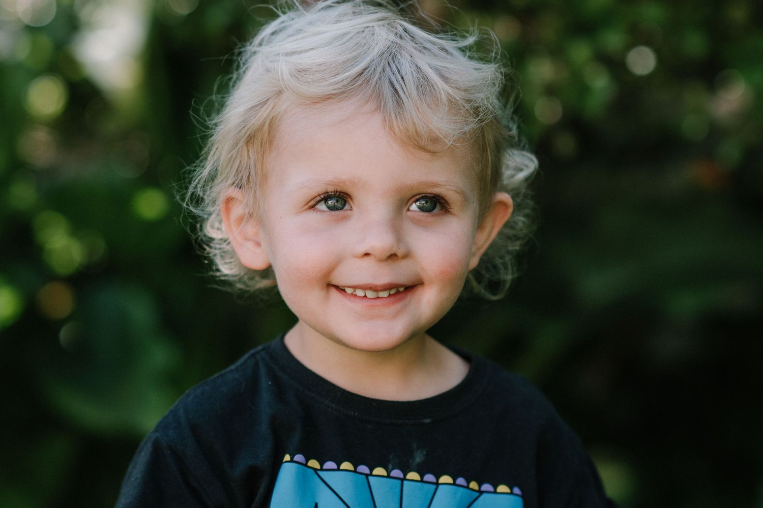 Young child with blonde curls smiles while wearing blue against a blurred natural background.