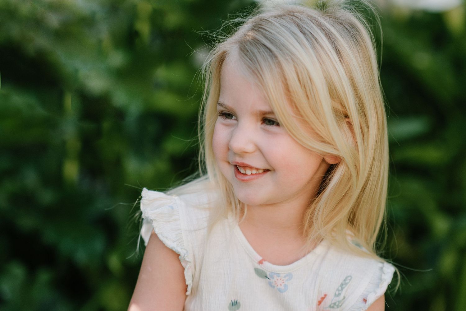 Child with blonde hair wearing white lace dress smiling outdoors with greenery in background.