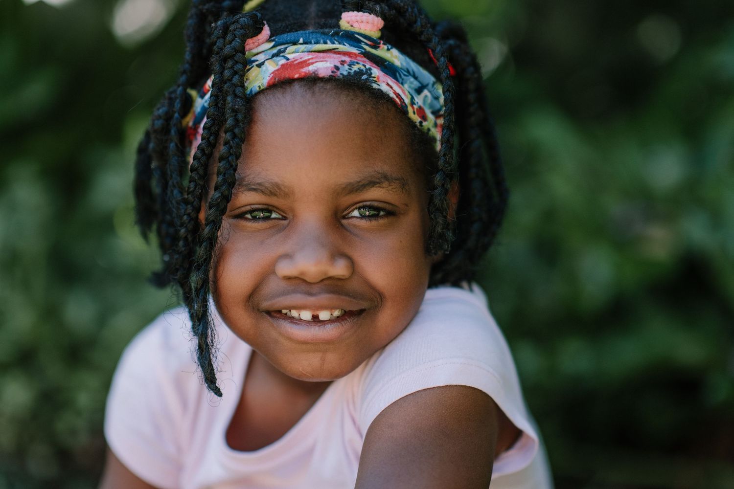 Portrait of a child with braids and colorful headband smiling brightly against green foliage.