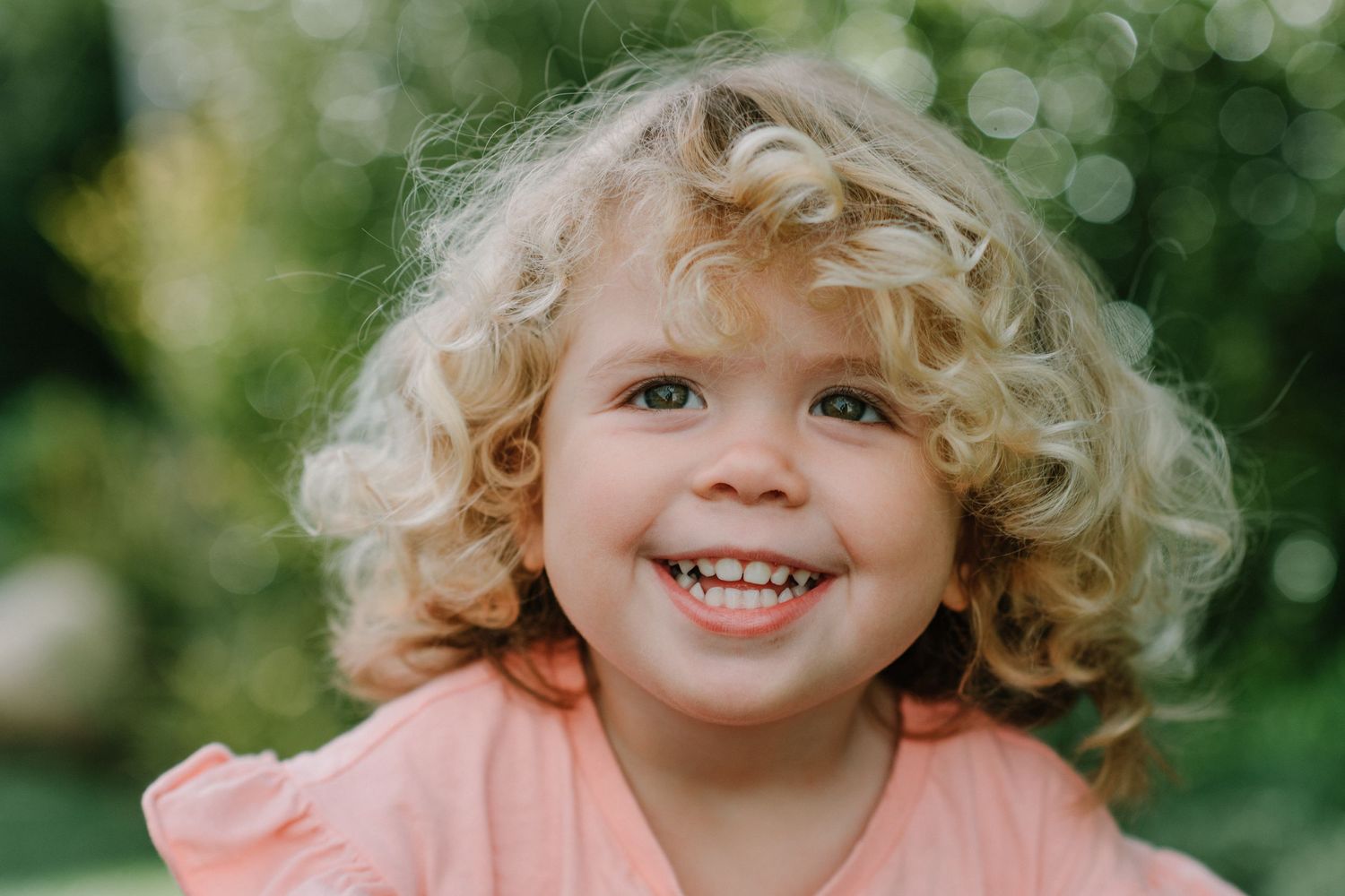 Child with blonde curls wearing coral top smiles against blurred green background.