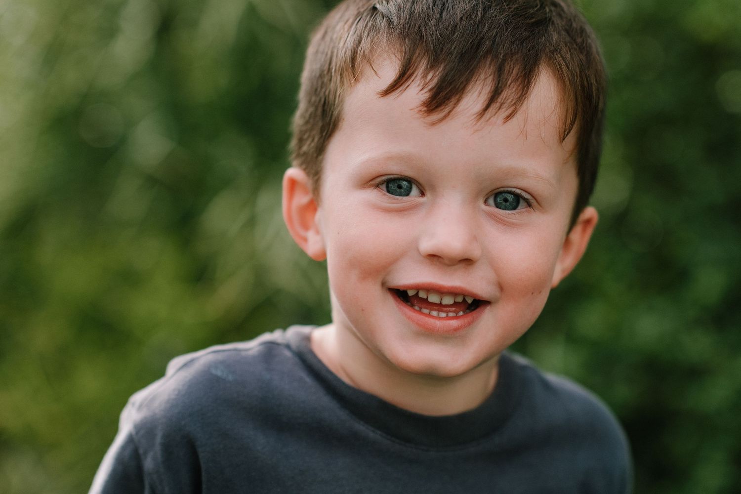 Child wearing dark shirt smiles brightly against natural green backdrop.