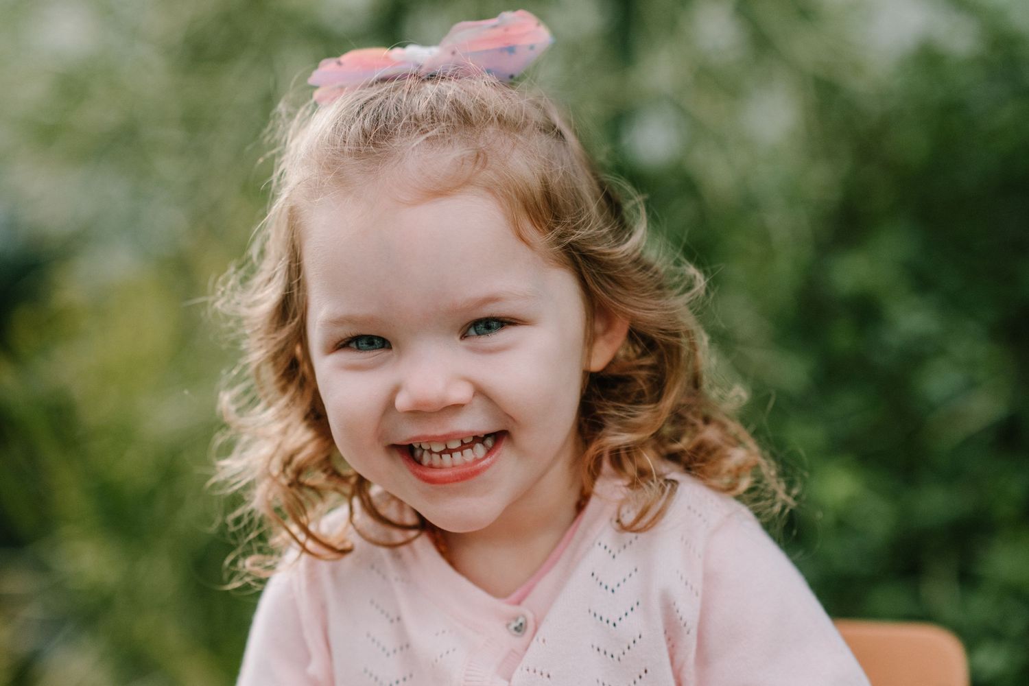 Child with curly hair wearing pink sweater smiles brightly against green background.
