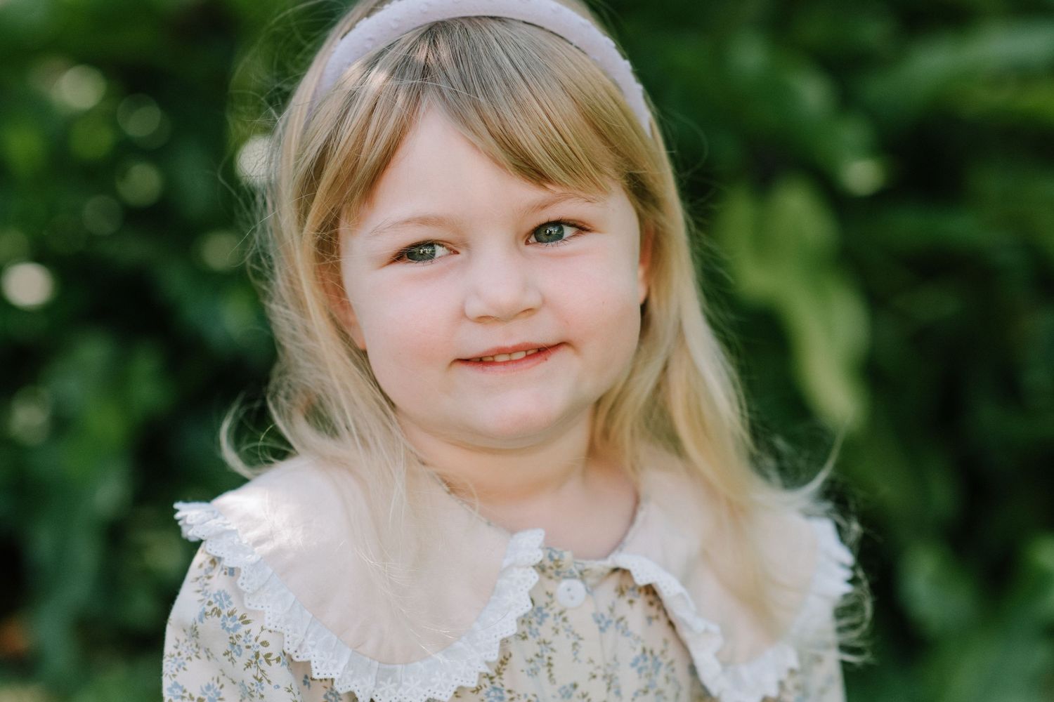 Child in white lace dress with blonde hair posing against blurred green background.