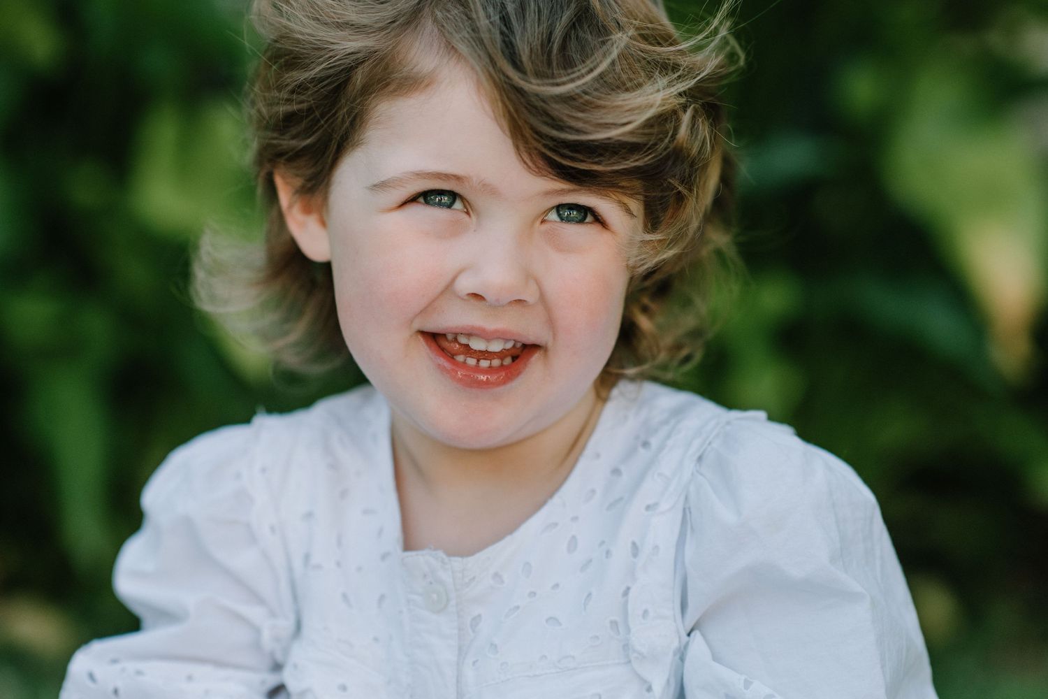 Child in white lace dress with curly hair laughing against green garden background.