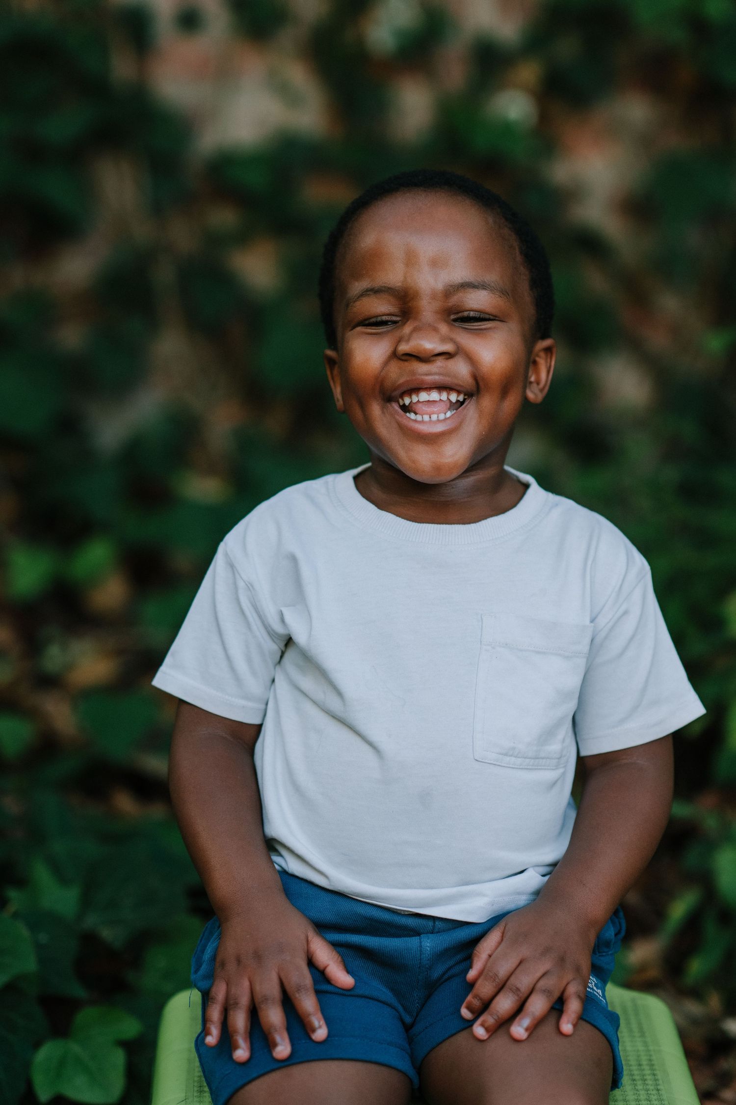 Child in white t-shirt and blue shorts sits outdoors with bright smile.
