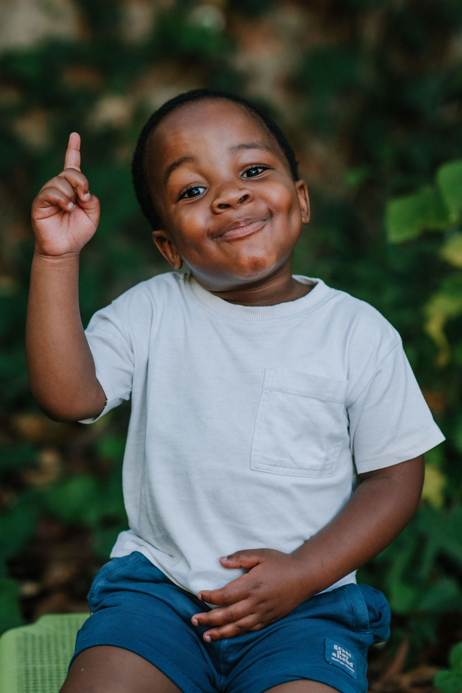 Young child in white t-shirt and blue pants showing joyful expressions against green foliage background.