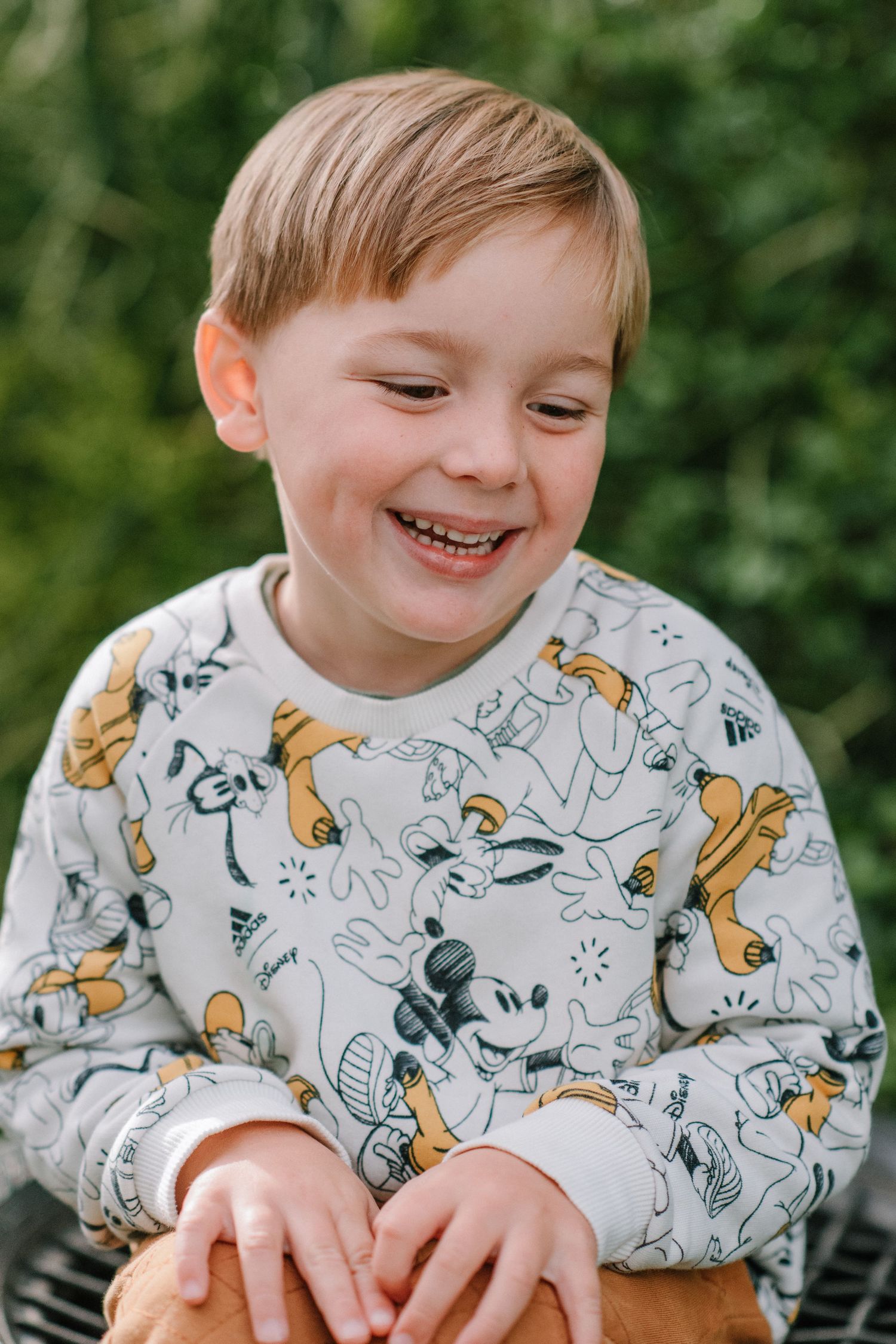 Young child wearing a Mickey Mouse print sweatshirt smiles while looking down outdoors.