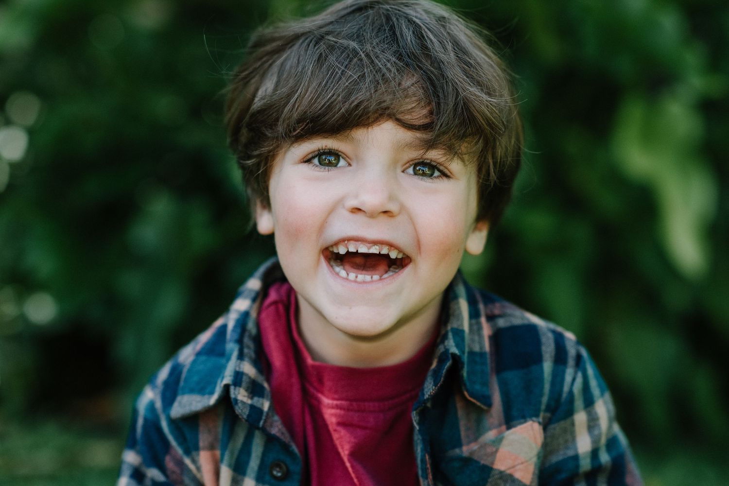 Child wearing plaid shirt and red top smiling outdoors with natural backdrop.
