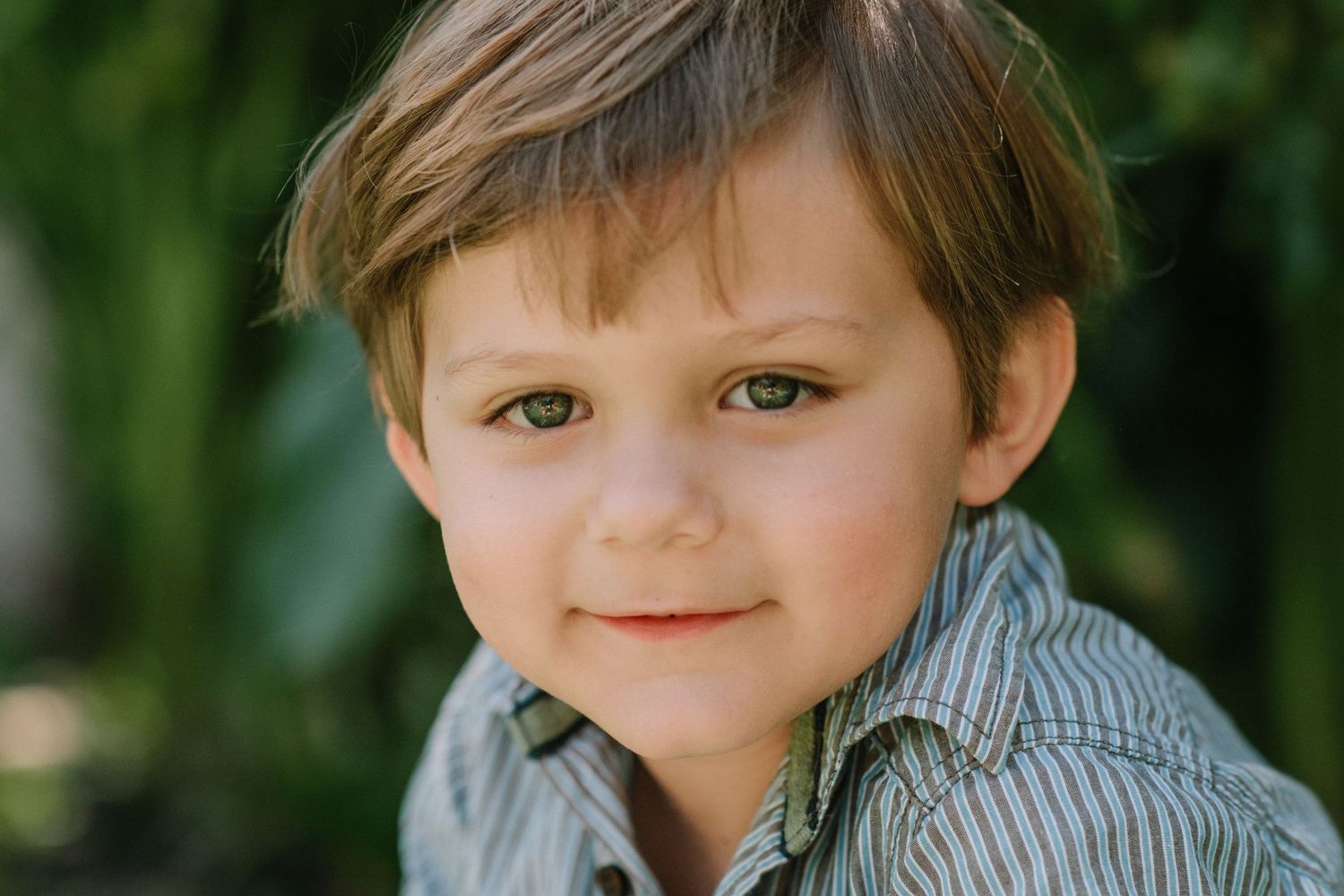 Child in striped shirt smiling warmly against green outdoor background.