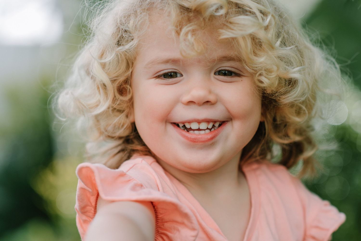 Child with curly blonde hair wearing peach-colored outfit smiles outdoors.
