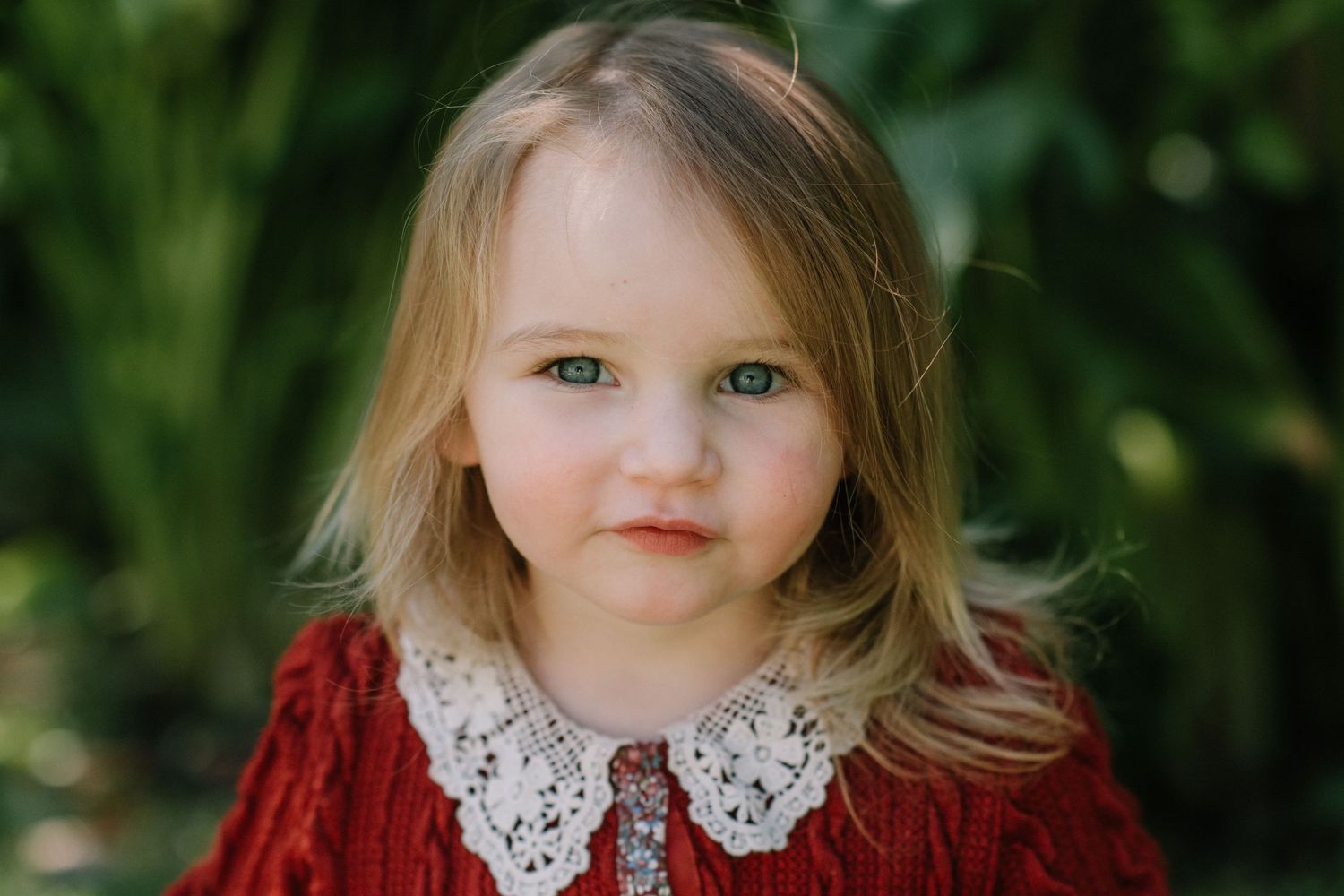 Child wearing a red dress with white lace collar against green garden backdrop.