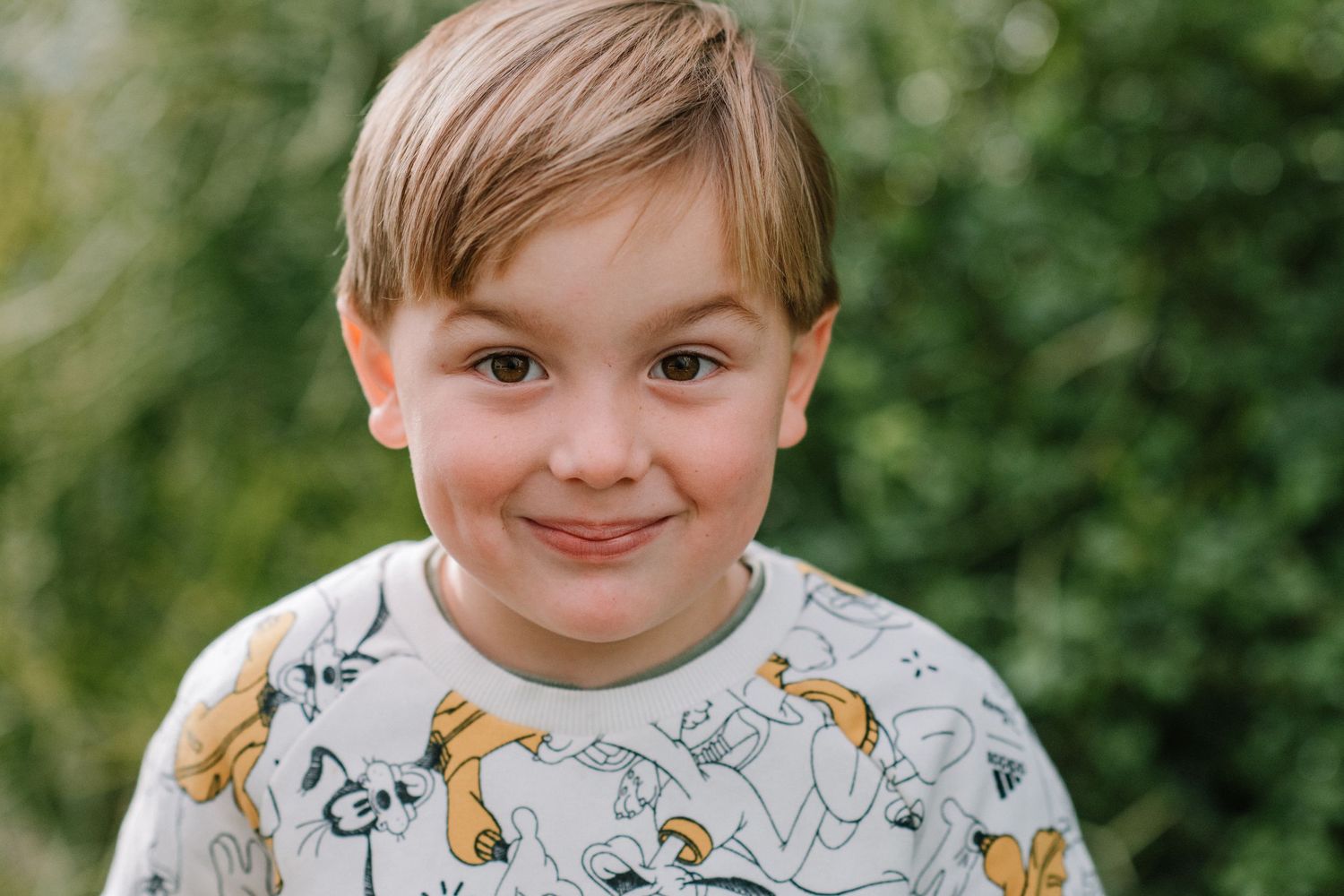 Child wearing cartoon print sweatshirt gives knowing smile outdoors.