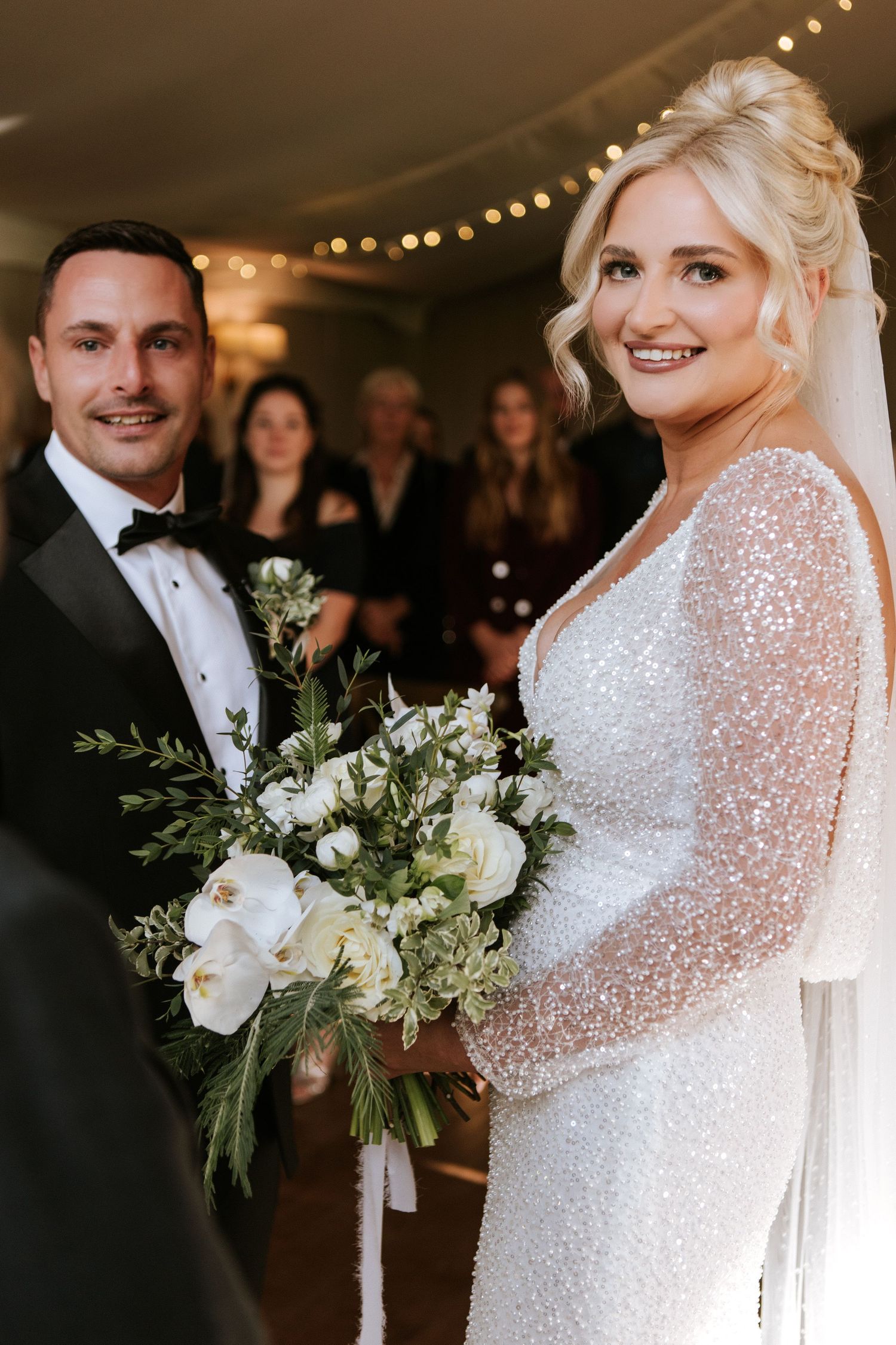 A bride in a sparkling long-sleeved wedding gown holds a lush white and green bouquet during an evening reception.