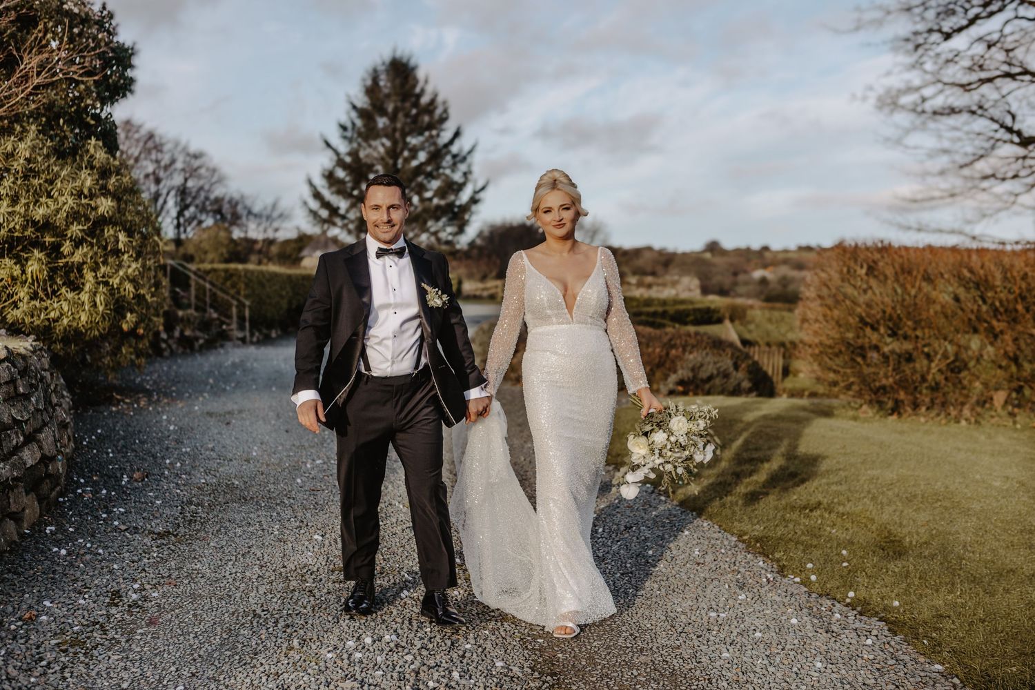 Wedding party walks along a gravel path surrounded by autumn trees and fields on a cloudy day.