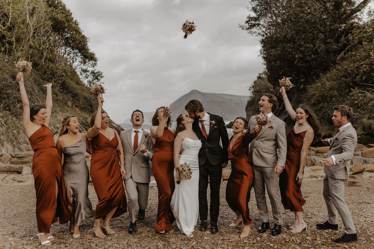 Wedding party in burnt orange and neutral attire celebrates joyfully on a scenic cliff overlooking the ocean.