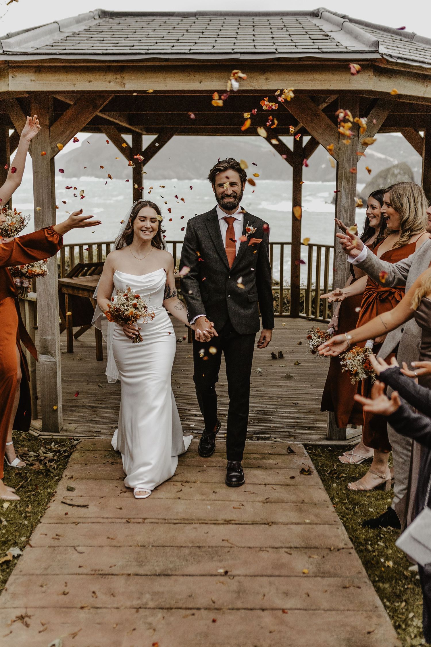 Newlyweds walk down wooden gazebo path while guests toss confetti in autumn wedding celebration.