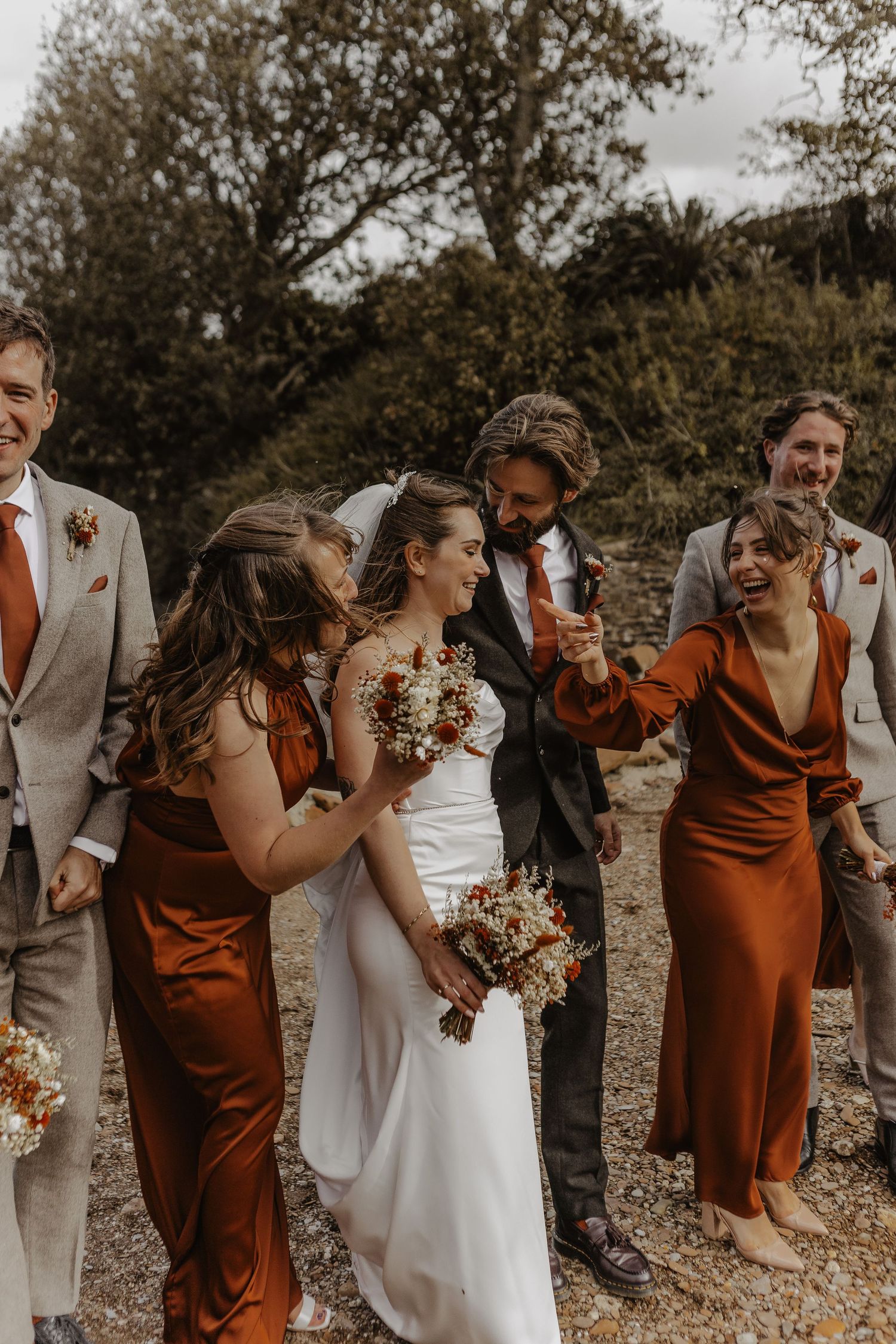 Wedding party in rust-colored attire shares candid laughing moment outdoors.
