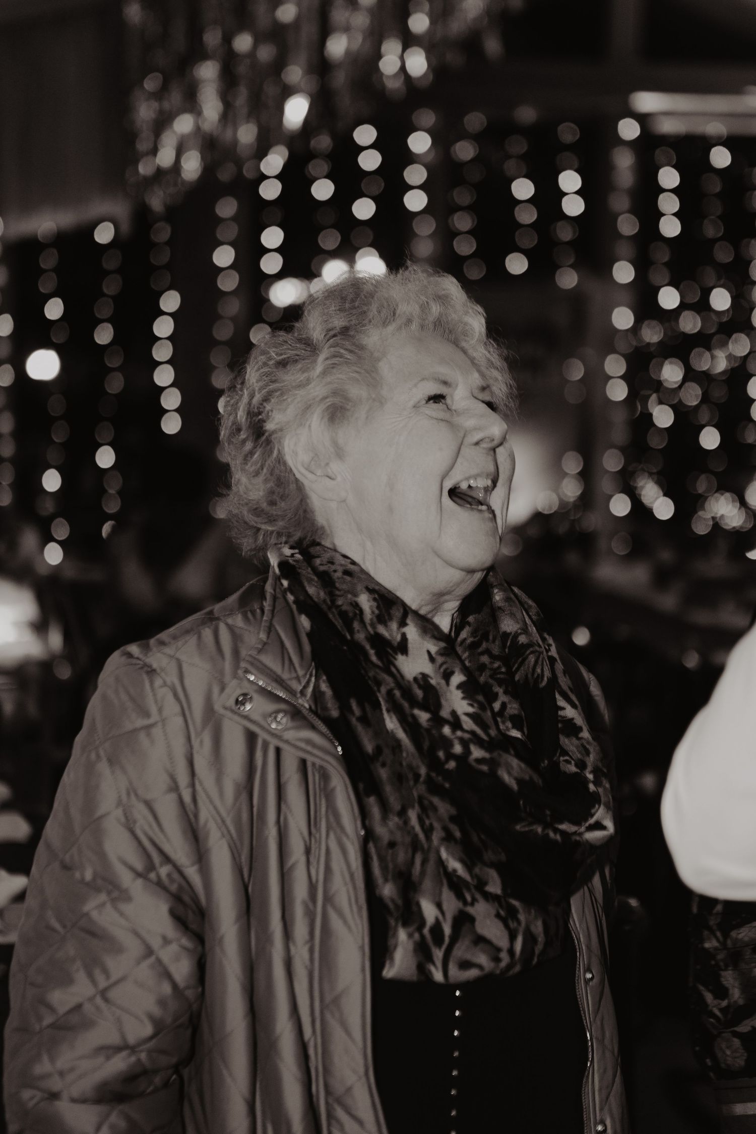 An elderly person in a winter coat and scarf stands against a backdrop of twinkling lights.