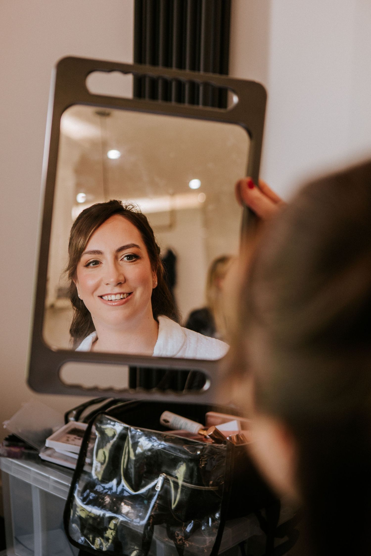 Bride smiling at her reflection in a handheld mirror during bridal makeup preparation session.