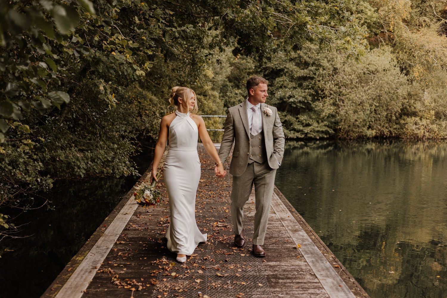 Bride in a white gown and groom in a gray suit walk hand in hand along a wooden pier beside a calm lake surrounded by lush green trees.