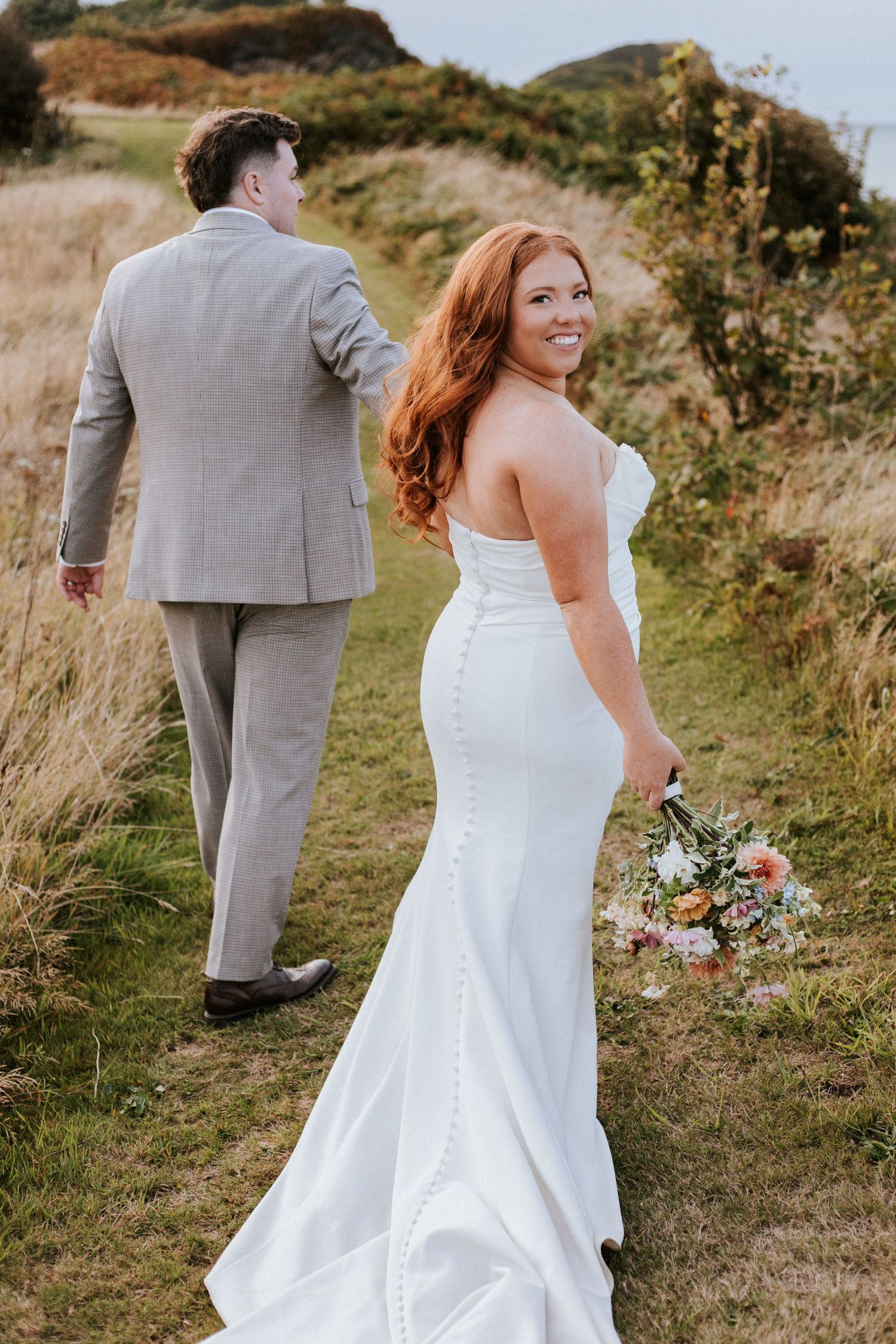 Newlywed couple walking together through scenic hillside meadow in gray suit and white wedding dress with bouquet.