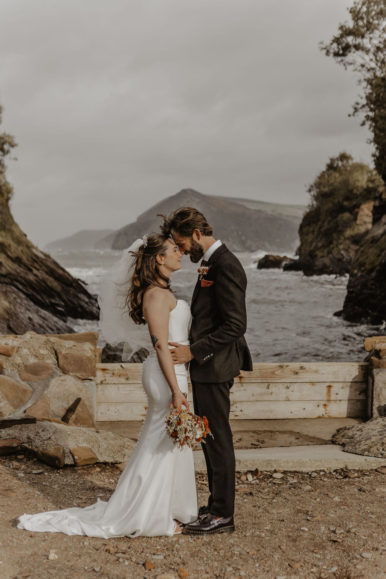 A couple shares an intimate moment on a rocky coastal shoreline with mountains and ocean in the background.