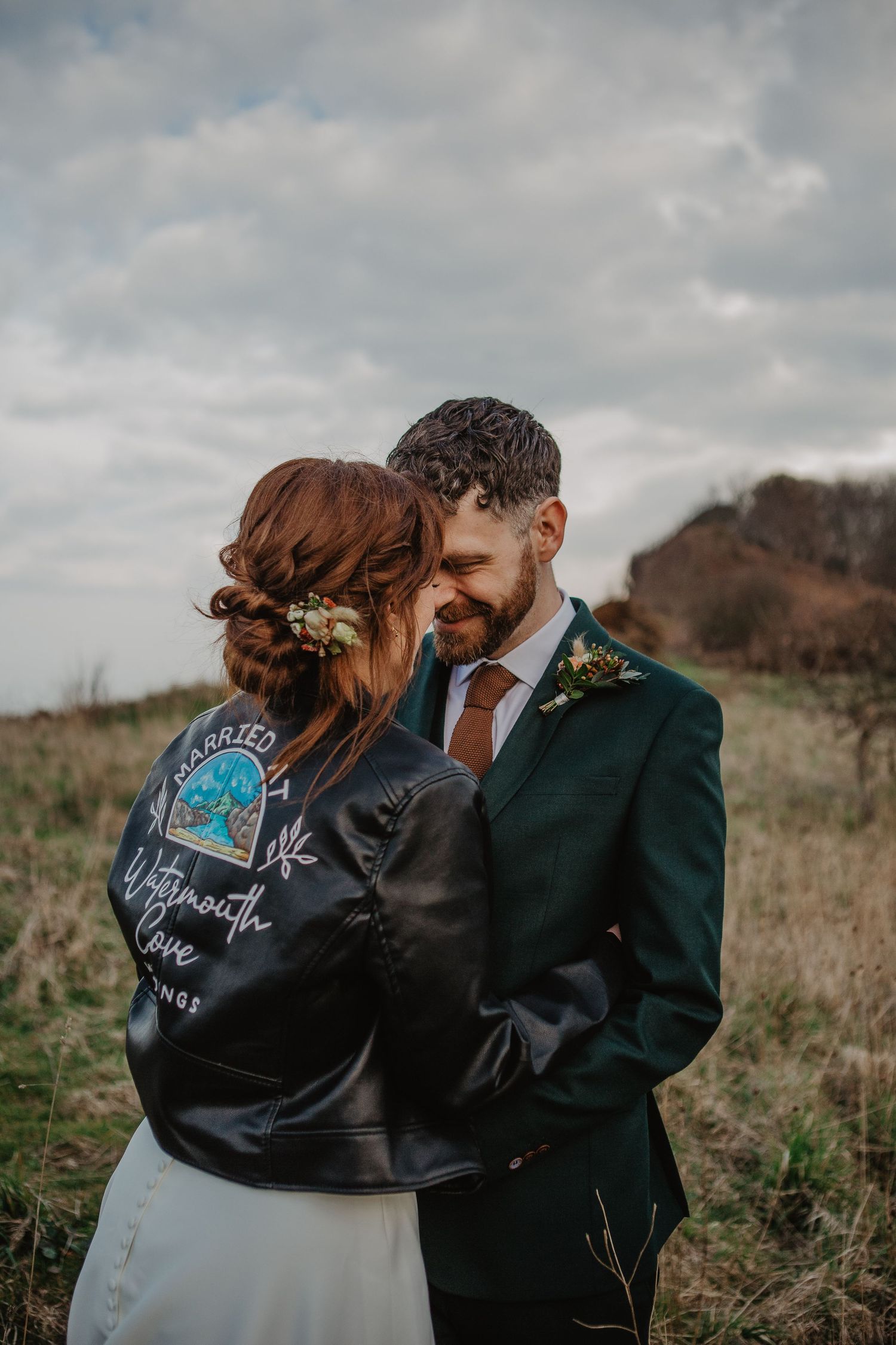 A couple shares an intimate moment outdoors while wearing a custom leather jacket with white handwriting on a cloudy day.