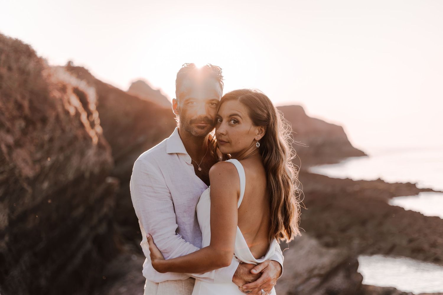 Couple embracing at golden hour on rocky coastal cliffs, woman in backless white dress, man in white shirt.