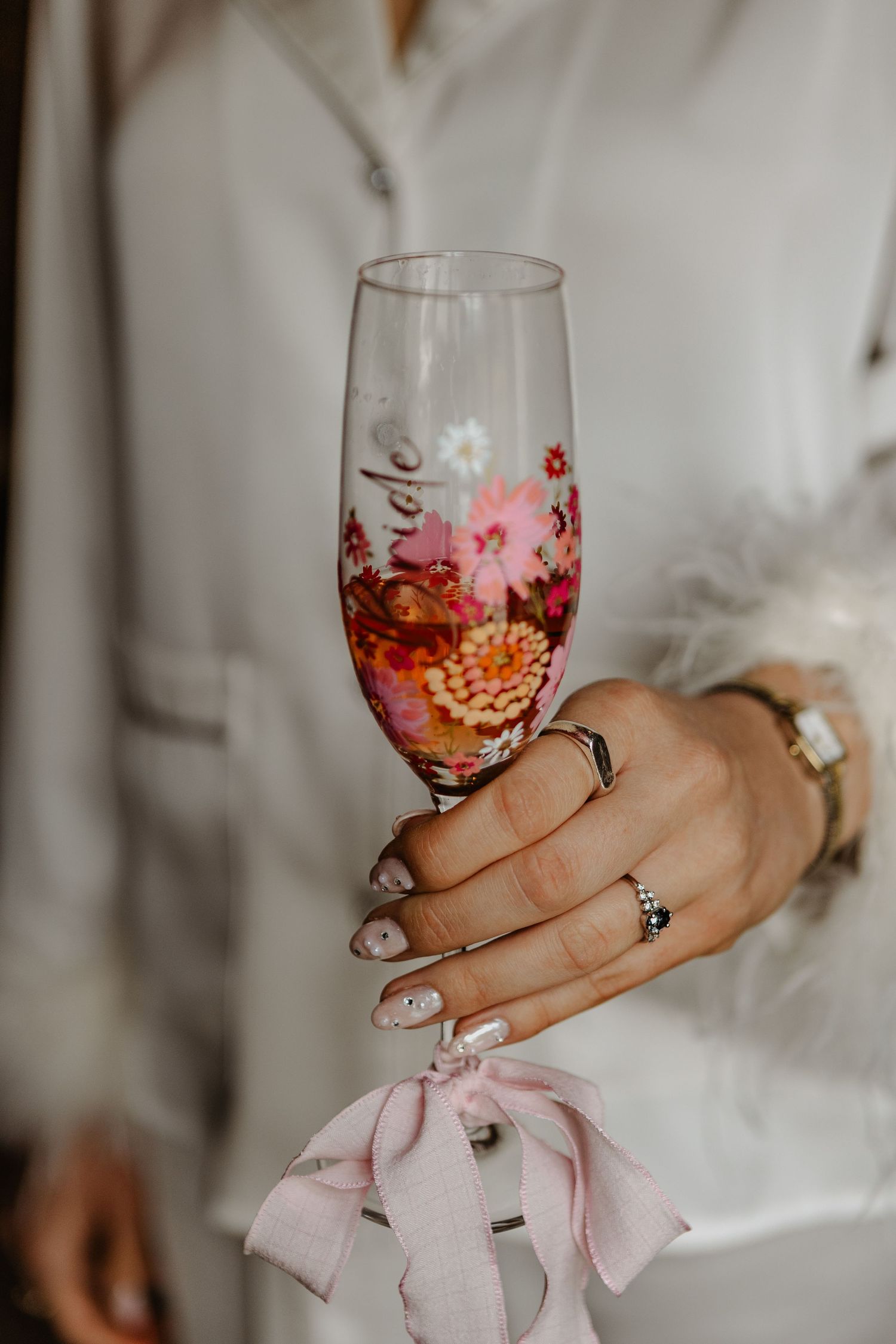 Hand holding a decorative wine glass with floral pattern while wearing delicate rings and pink bow detail.