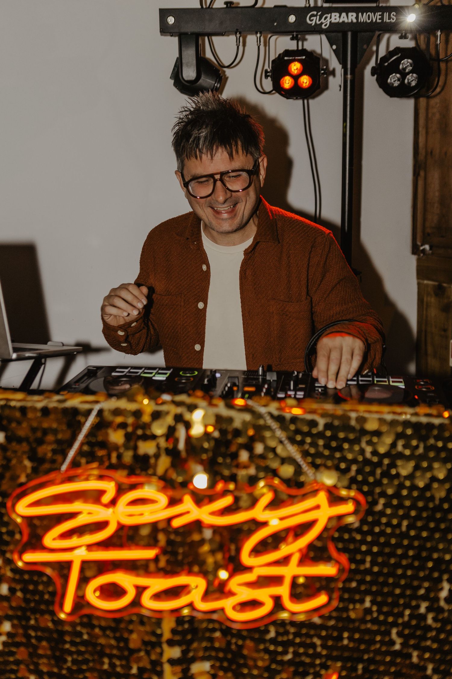 DJ performs at wedding reception behind illuminated 'Spicy Toast' neon sign.