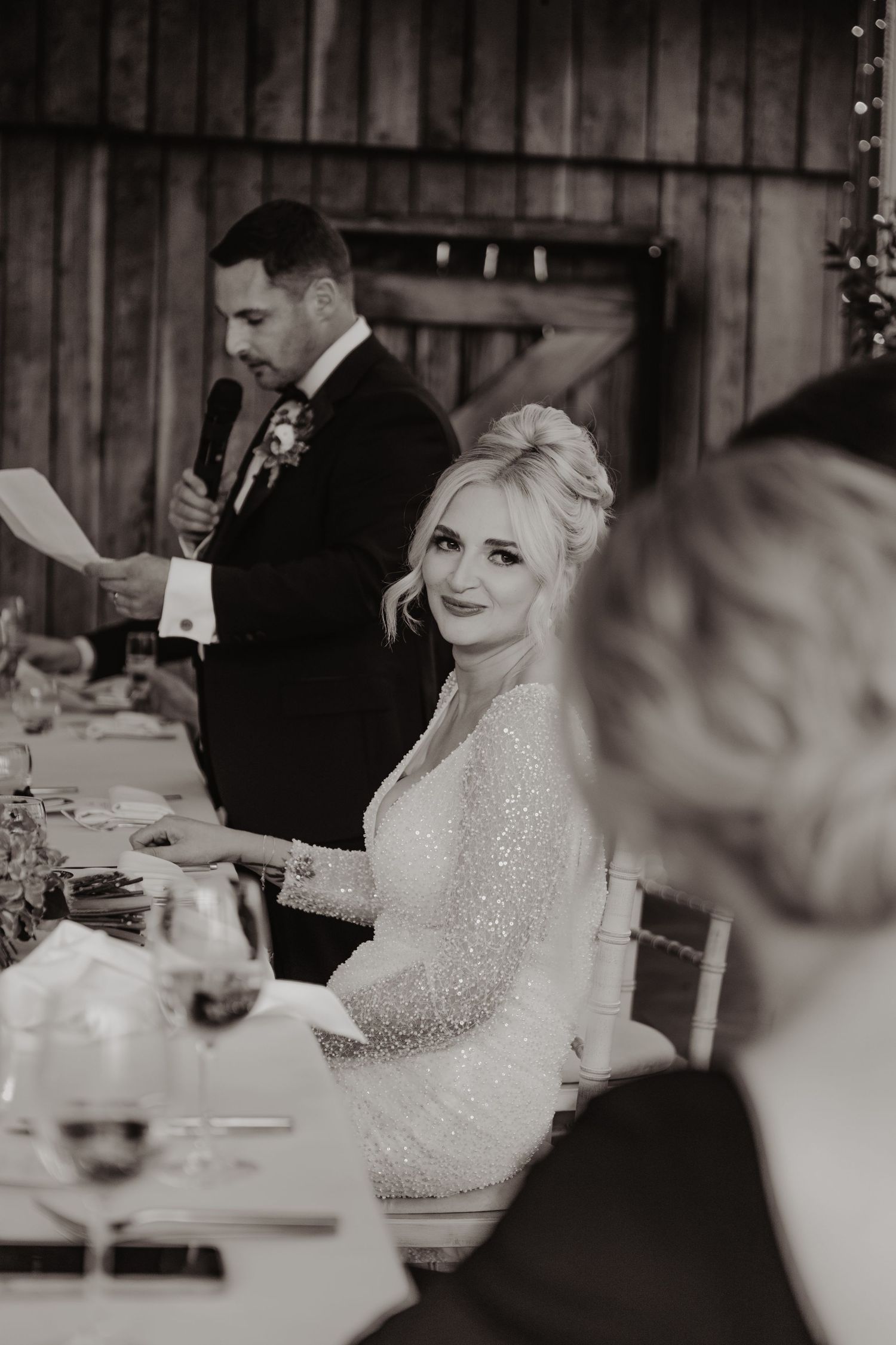 Wedding reception guests seated at tables in a rustic barn venue with twinkling lights overhead.